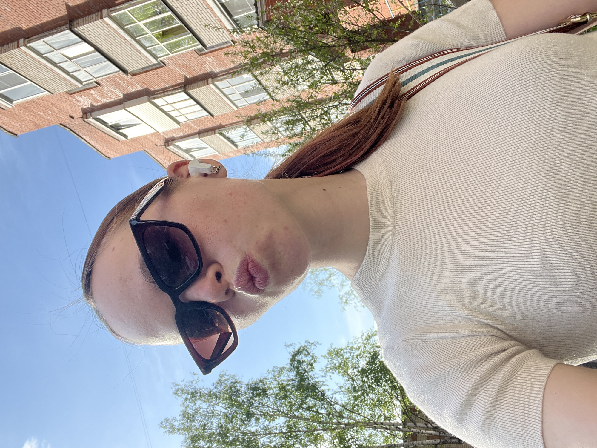 A medium-angle photograph depicts a woman looking down at the camera, with elements of the urban environment visible in the background. The woman has light skin, light brown hair, and wears dark sunglasses and a light-colored, textured shirt with a striped strap across her shoulder. She is wearing an earpiece in her left ear.
Behind the woman, the sky is visible along with the tops of trees. A brick building with a repeating pattern of square windows appears at the top left of the image. The location is Chaikovsky, Russia, inferred from metadata not shown.