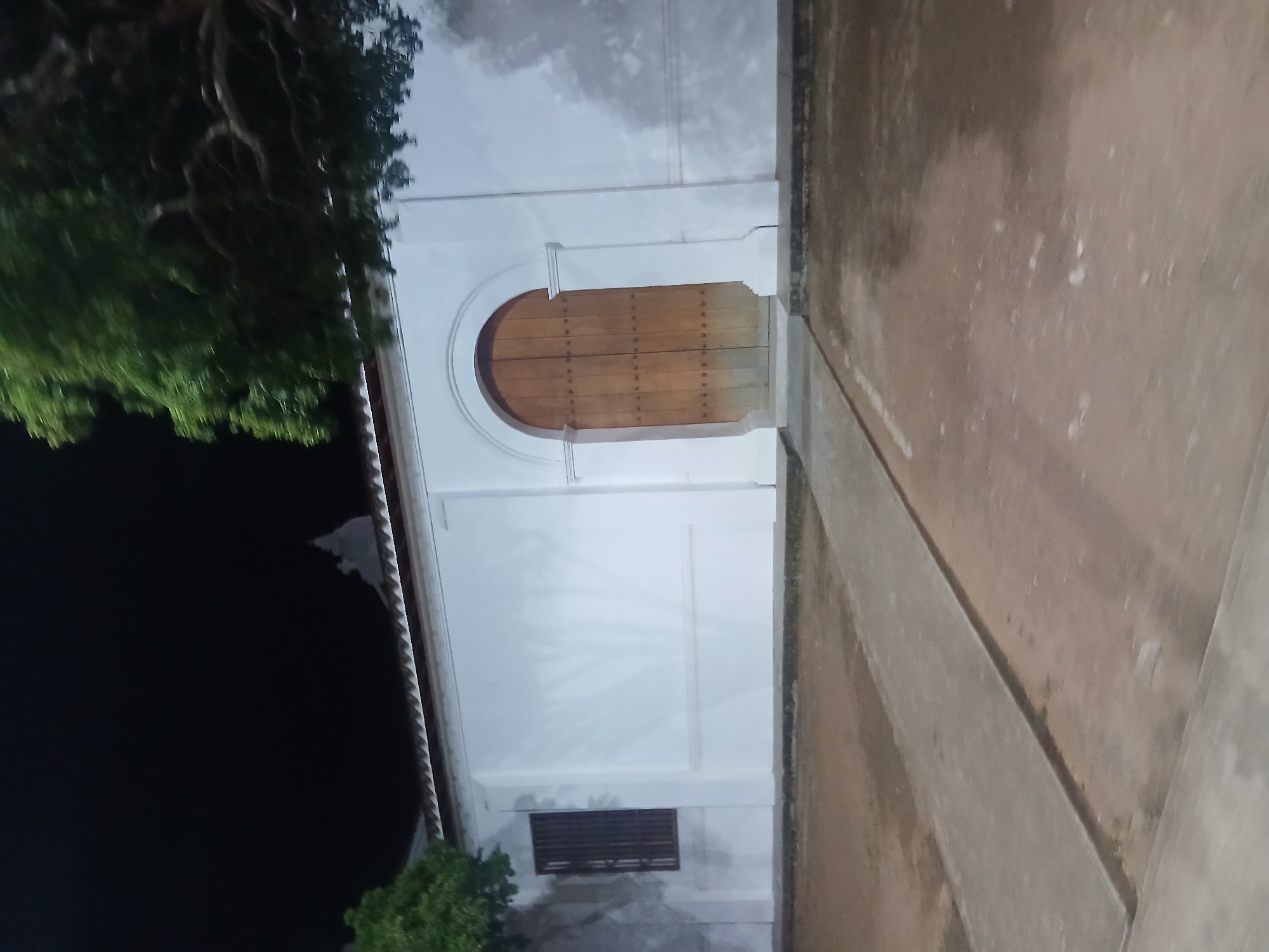 The image shows a building with a white facade and a large arched wooden door at night. A paved walkway leads to the door. There is a window with vertical bars on the left side of the building. Trees are visible on the left and right sides of the building. The ground in front of the building is dirt. The image appears to have been taken in Santa Ana, Venezuela.