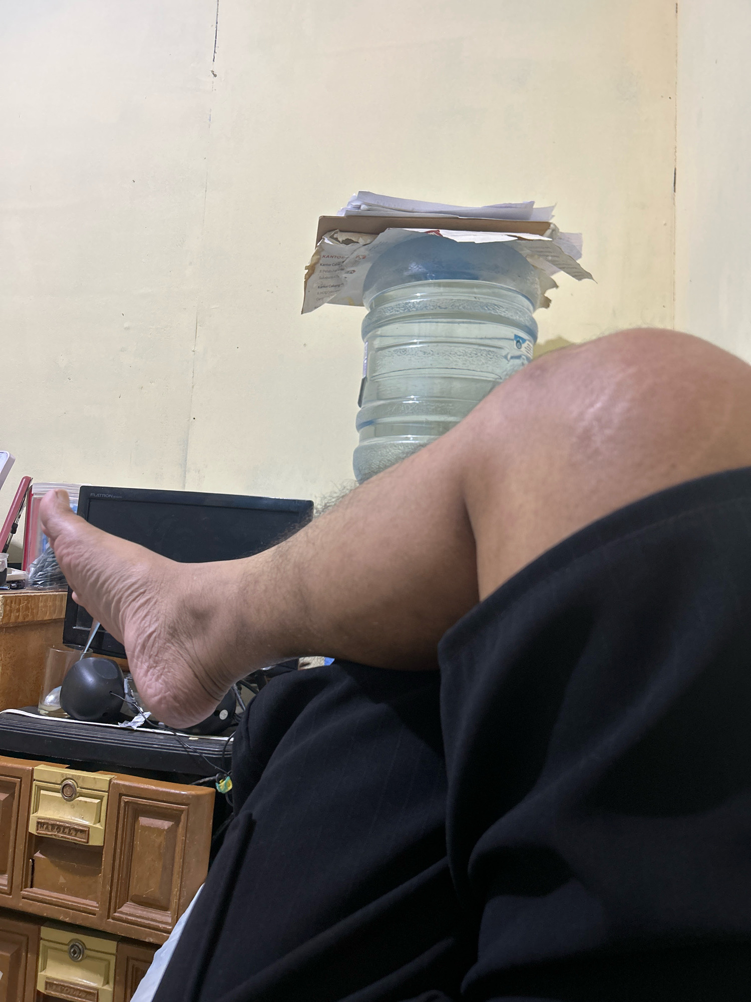 The image shows a close-up view of a person's bare legs and feet resting on what appears to be a desk. The individual is wearing black shorts. In the background, a large water cooler jug is visible, stacked with papers. A computer monitor and drawers with labels are also present. The setting is indoors, with a plain, light-colored wall behind the desk. The overall impression is of a casual, possibly home office environment. The scene is located in Sukanagara, Indonesia.