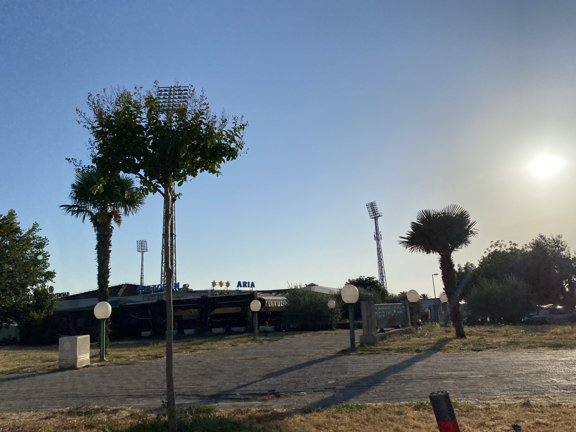 The image depicts an outdoor scene at what appears to be a restaurant or establishment in Bar, Montenegro, under a clear blue sky with the sun shining brightly in the upper right quadrant. In the foreground, a paved walkway or driveway is visible, with dry grass and vegetation bordering it. Several light fixtures with round globes are placed along the walkway.
In the middle ground, a building with an awning and signage that reads "*** ARIA" is set back from the walkway. A tall, slender palm tree stands to the left of the building, and another, shorter palm tree is positioned to its right. Behind the building, there are several trees and bushes. Tall, lattice-like light towers, possibly for a sports field, are visible in the background. One tower is prominent behind the left side of the building, and another is further to the right.
To the right of the building, a sign reads "RESTAURANT" with an arrow pointing to the right. The overall impression is of a somewhat dated or perhaps off-season establishment in a sunny, coastal environment. No people are visible in the image. The scene is captured during daylight, with the sun casting long shadows.