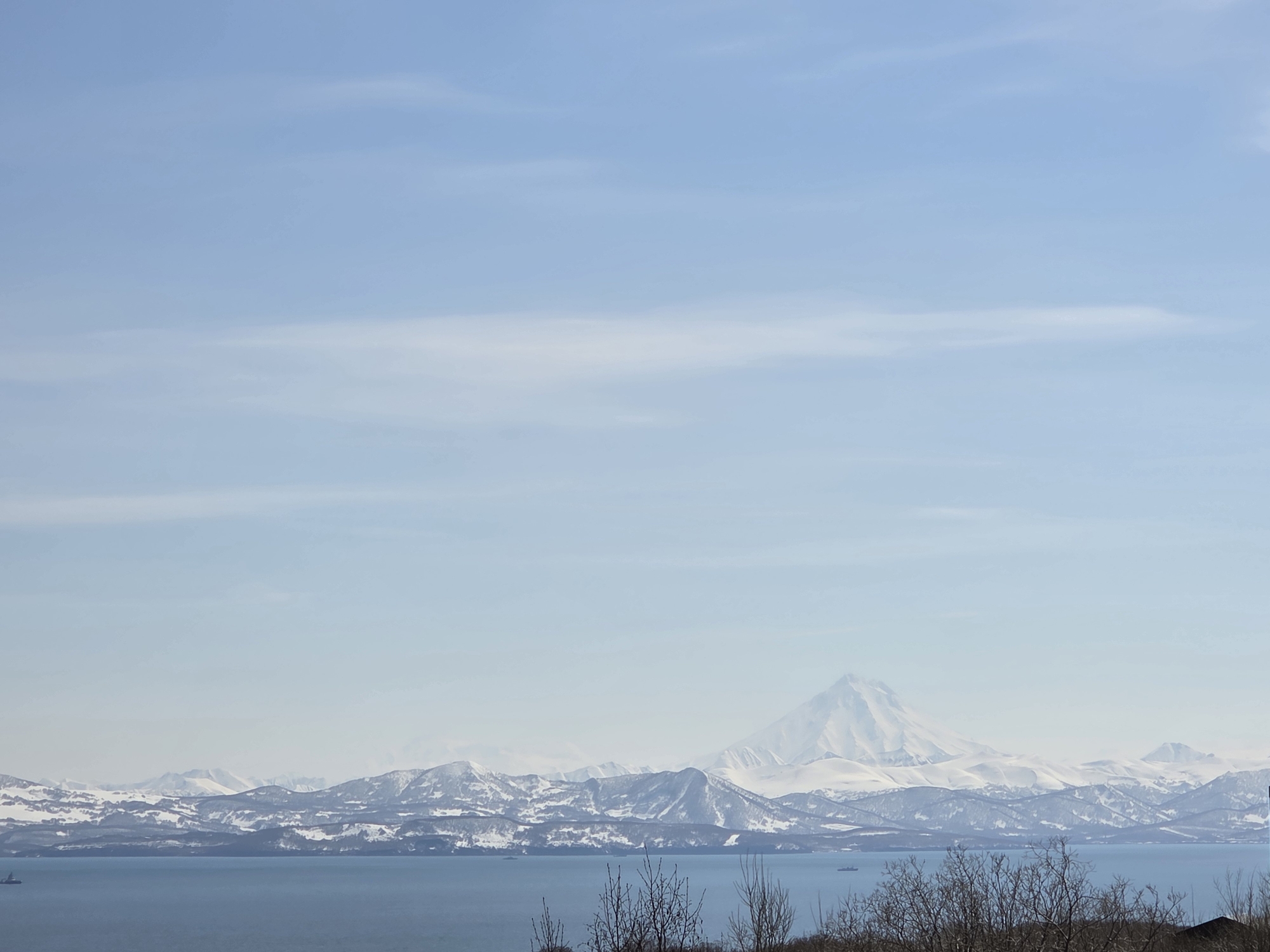 The image presents a landscape featuring a body of water in the foreground, likely a bay or sea, bordered by land with sparse vegetation consisting of thin trees. In the background, a mountain range is visible, partially covered in snow, with one prominent conical peak. The sky is light blue with faint clouds. The location is unknown, as it is not specified in the prompt.