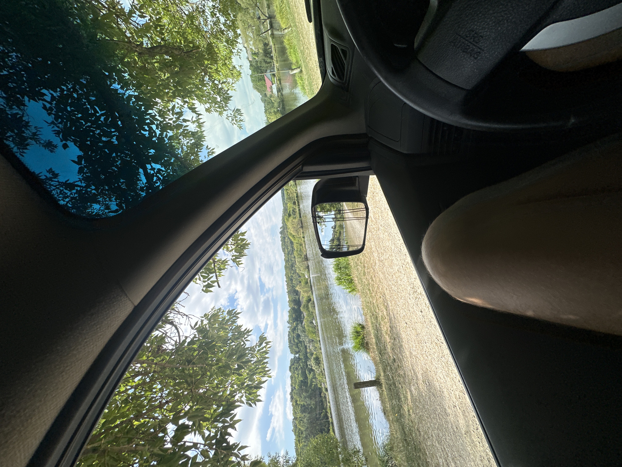 The image is a view from the inside of a car, looking out the driver's side window and through the windshield. A person's leg is visible in the foreground on the right side of the frame. The car is parked on a gravelly area next to a body of water, likely a river or lake, with trees and rolling hills in the background. The sky is blue with scattered clouds. In the distance, across the water, several houses and trees are visible on the hillside. The scene appears to be a rural or natural setting. There is no indication in the image that places it specifically in Makiv, Ukraine.