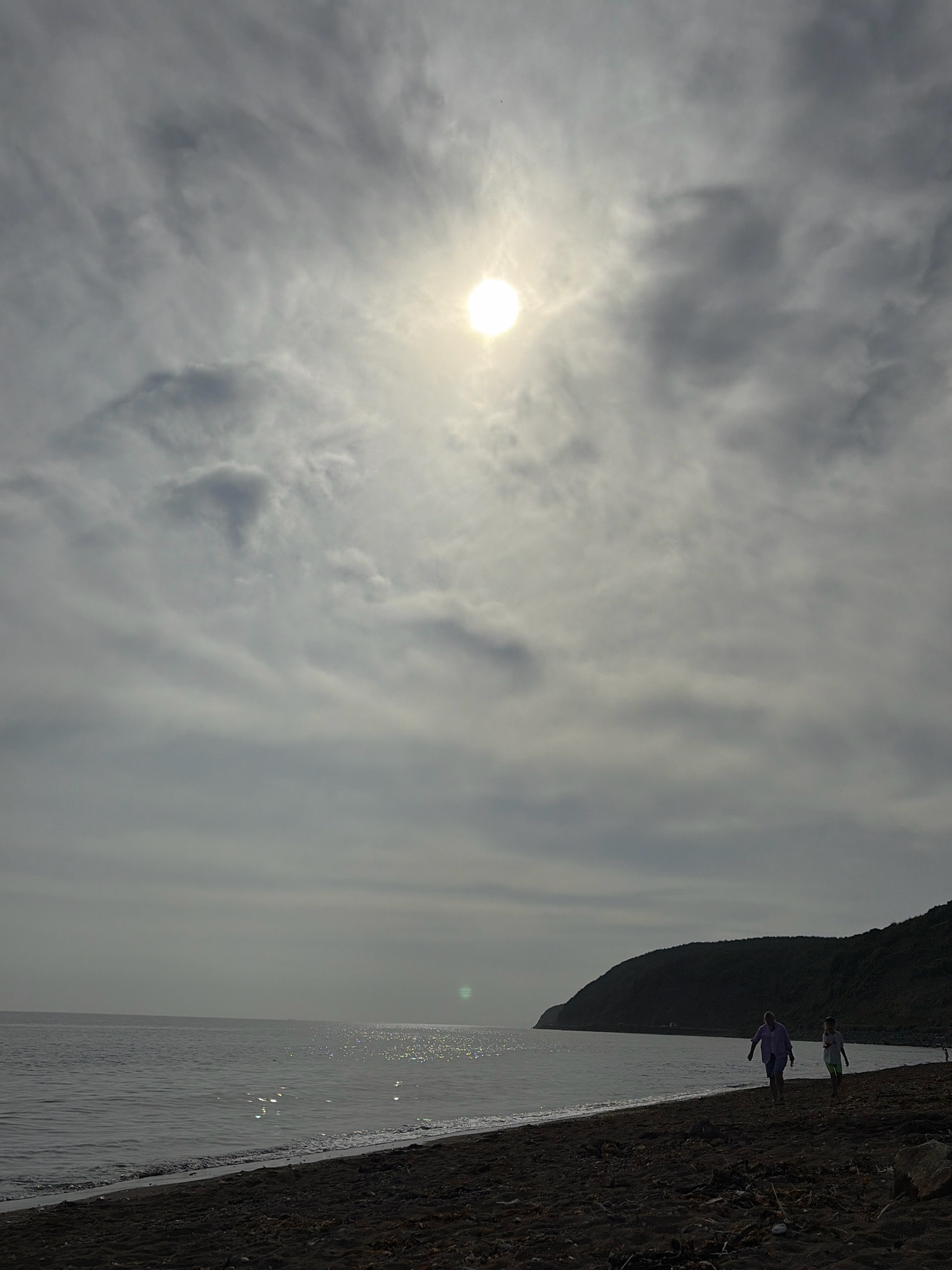 An overcast sky with the sun partially visible through the clouds dominates the upper portion of the image. The sun is a bright, blown-out circle. Below the sky, the scene opens to a dark, sandy beach. The sea is calm, with small waves lapping at the shore. In the middle ground, two people are walking along the beach. One person is wearing a light purple shirt and dark shorts, and the other is wearing a white shirt and light-colored shorts. They are walking away from the camera, towards a large, dark hill that forms the background on the right side of the image. The overall lighting suggests it is daytime, possibly late afternoon or early morning, given the low angle of the sun and the muted tones. The location is the coast of Korsakov, Russia, indicated by the distinctive dark sand beach and the presence of a hill on the coastline, common features in the area.