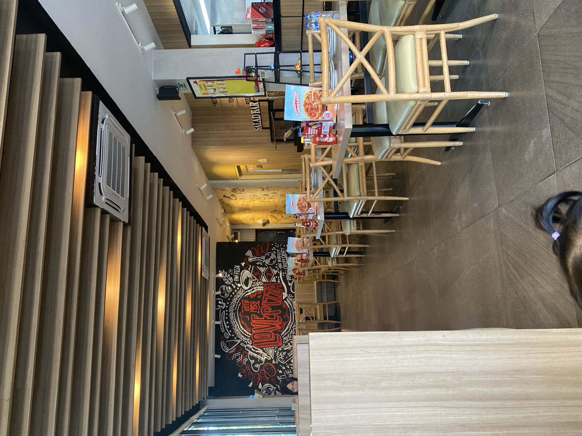 The image shows the interior of a pizza restaurant, likely a Pizza Hut, in Watampone, Indonesia. The restaurant features a modern design with a wooden slatted ceiling and a black wall with a red and white mural that says "LOVE PIZZA ESTD 1958". The dining area is furnished with tables and chairs, with some tables set for customers. A "SALAD BAR" sign is visible on the wall. The floor is tiled in a dark brown color. There are several bottles of ketchup and mustard on the tables, along with menus. In the foreground, a child's head with a purple hair tie is visible, suggesting a family dining experience. A woman is partially visible behind a wooden partition to the left of the image. The restaurant appears to be clean and well-lit, with an air conditioning unit mounted on the ceiling. The overall atmosphere suggests a casual dining establishment.