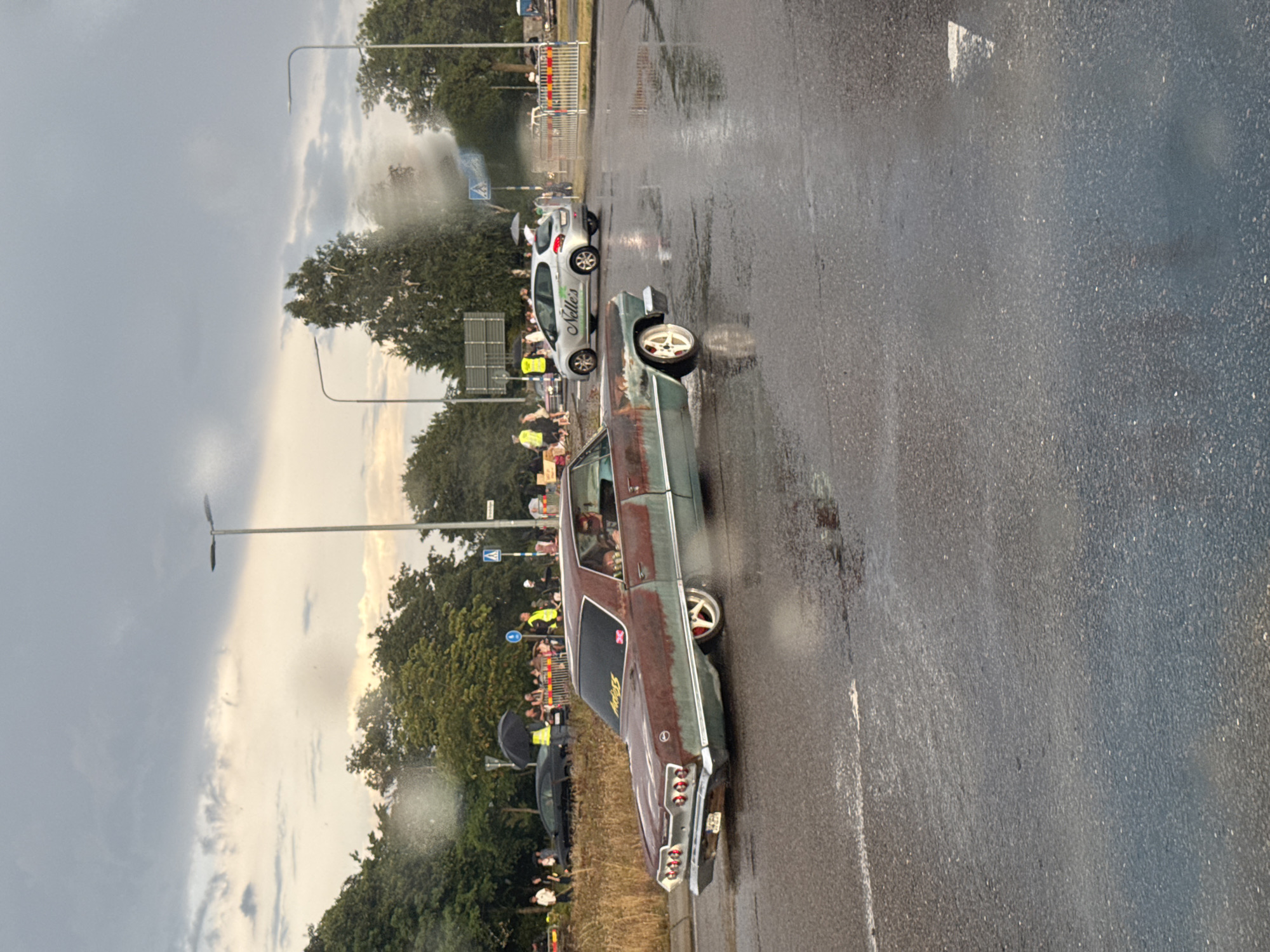 In Falkenberg, Sweden, a rusted and weathered light blue Chevrolet Impala is seen driving on a wet asphalt road. The car has custom red and white wheels. A person is visible in the driver's seat. Behind the Impala, a silver Peugeot 208 with the text "Nelle's" on its side is also on the road. There are several people gathered on the side of the road, some holding umbrellas, suggesting it may be raining or has recently rained. A black car is parked to the left. The environment appears to be a street with trees and streetlights. The sky is cloudy with hints of sunlight.