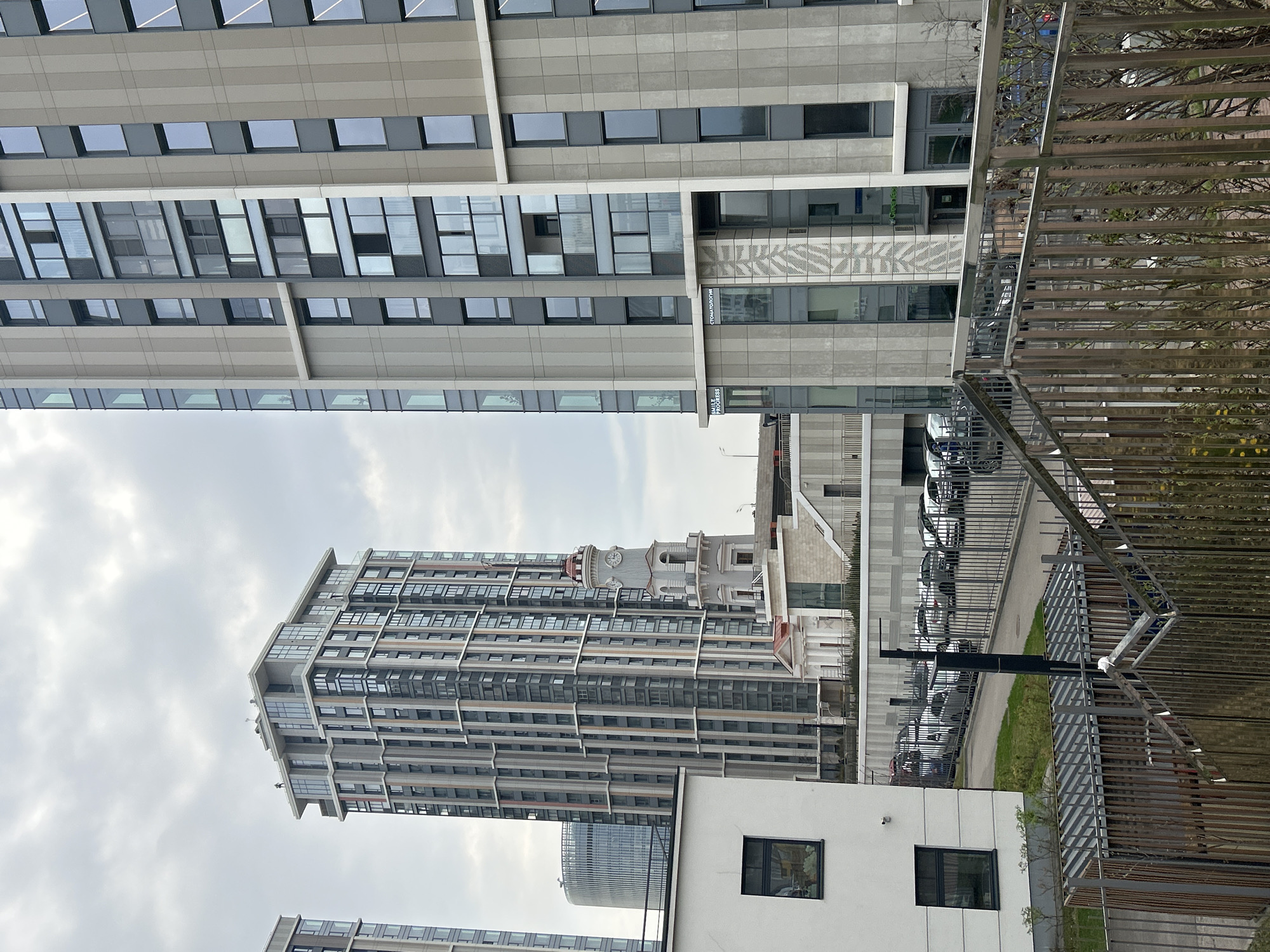 This media file captures an urban street scene in Moscow, Russia, characterized by a mix of modern and older architecture under an overcast sky.
The scene is dominated by tall, contemporary buildings. On the left, a prominent high-rise features a facade of numerous windows and light-grey panels, reflecting a sleek, modern design. Another contemporary building, possibly part of the same complex, stands in the right foreground. It has similar architectural elements, including large windows and grey paneling.
In the mid-ground, a distinctive building with an ornate clock tower stands out, providing a striking contrast to the surrounding modern high-rises. This structure appears to be older, showcasing more traditional architectural details and a domed top. Several other modern buildings are visible further in the background.
A street runs through the scene, with multiple cars, mostly light-colored, parked along its edge or in a designated parking area. A metal fence or barrier with a slatted design occupies a significant portion of the right foreground, partially obscuring the view and suggesting a property boundary or walkway. Sparse, bare tree branches are visible through this fence.
The weather is overcast, with a grey, cloudy sky providing soft, diffused light across the scene. No specific activities or people are discernible, presenting a calm, everyday urban snapshot.
Visible text on one of the modern buildings includes "СТОМАТОЛОГИЯ" (Stomatologiya), which translates to "Dentistry" or "Dental Clinic," and "SMILE PROGRESS," likely the name of the clinic.