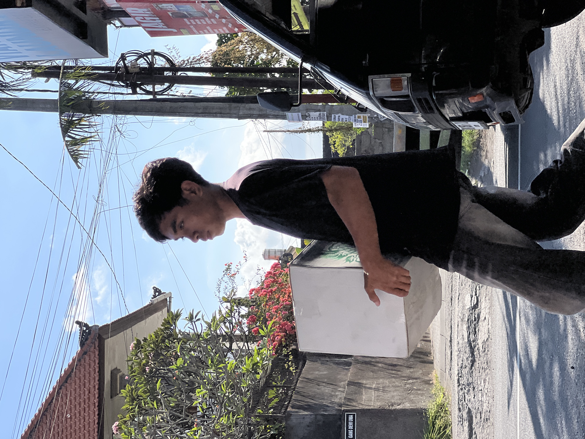 A young man walks down a cracked street in Denpasar, Indonesia, carrying a large white box. He is wearing a black t-shirt and grey jeans. To his left, a wall with a sign reading "GANG DEWI UMA" is visible, along with a fence and lush green foliage with pink flowers. To his right, a black van is parked, and overhead, a tangle of electrical wires hangs against a bright blue sky with scattered clouds. The scene appears to be on a sunny day, with shadows cast by the man and the van.