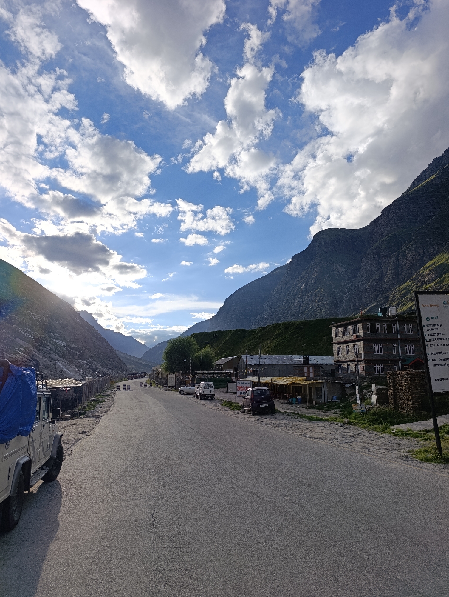 A long asphalt road cuts through a valley with steep, rocky mountains on either side. The sky above is a vibrant blue, dotted with dramatic white clouds that cast shadows on the landscape. On the right side of the road, a multi-story building constructed of stone with white-framed windows stands prominently. In front of this building, several smaller structures, likely shops or accommodations, are visible with yellow awnings. To the left of the road, a white vehicle, possibly a jeep with its cargo covered by a blue tarp, is parked. Further down the road, other vehicles and a cluster of buildings are scattered along the valley floor, suggesting a small settlement or roadside stop in Damphug, India. A sign with text in Hindi is visible near the building on the right. The overall impression is of a remote, mountainous region bathed in the soft light of what appears to be late afternoon or early morning.