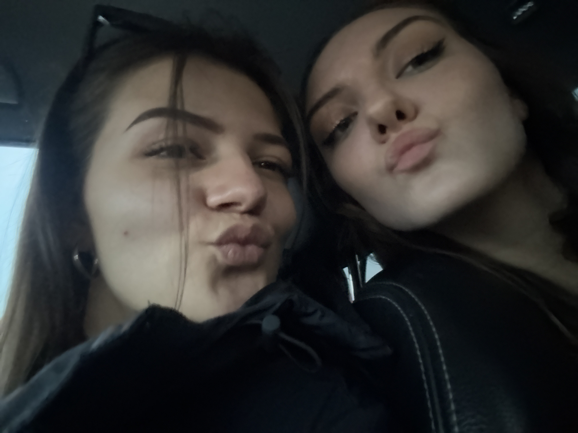 Two young women are posing for a selfie in what appears to be the interior of a car. They are close together, both making a "kissing" or "pouting" facial expression.
The woman on the left has straight, dark hair and her sunglasses are pushed up on her head. She has a mole on her face. She is wearing a dark-colored jacket with a drawstring. She is wearing small hoop earrings.
The woman on the right has dark hair and makeup, including eyeliner. She is wearing a dark-colored jacket with a white stitching detail.
The background is blurry and dark, suggesting the photo was taken in a car with dark upholstery.
The location of the photo is unspecified and can not be determined from this image. Therefore, the location in the city of Sofia, Bulgaria, is indeterminate.