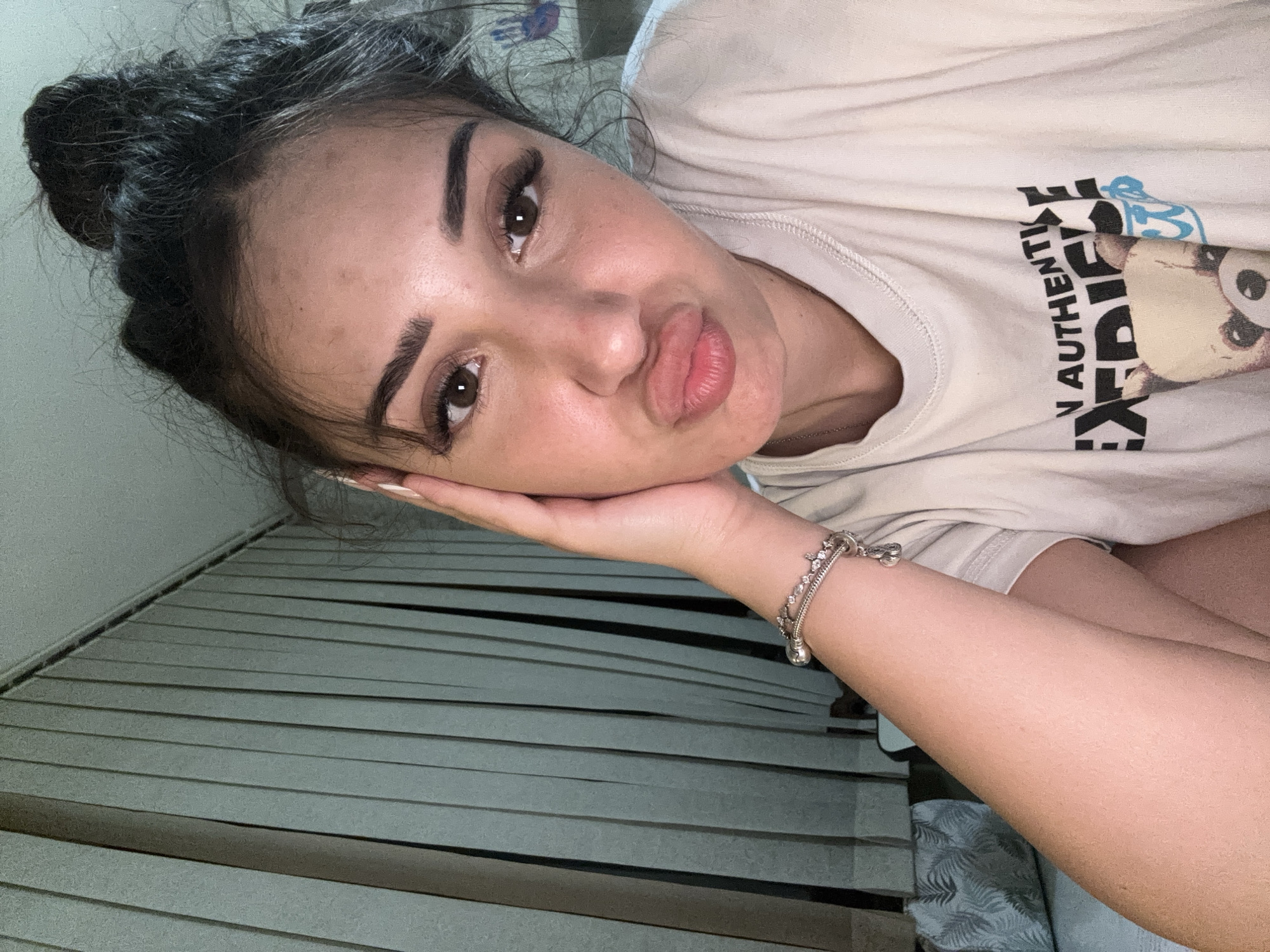 A young woman with dark hair pulled up in a messy bun and dramatic false eyelashes is leaning her head on her hand, puckering her lips. She is wearing a light-colored t-shirt with a graphic print of a bear and text that reads "AUTHENTIC" and "EXPERIENCE." A silver bracelet adorns her wrist. The background consists of vertical blinds. The image is a close-up selfie, suggesting it was taken indoors, likely in a personal space like a bedroom or living room. No specific details about the city of Plovdiv, Bulgaria, are discernible from the image itself.
