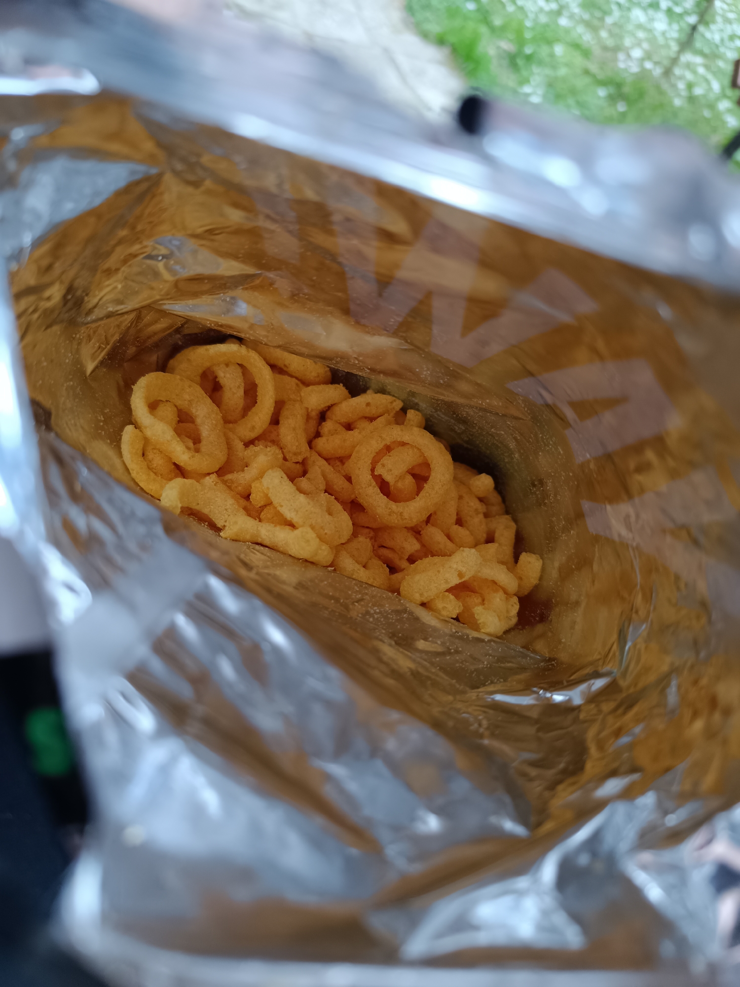The image provides an overhead view looking down into a partially consumed bag of snacks, likely taken in Pleven, Bulgaria. The main subject is an open foil or metallic-lined snack bag, with a modest quantity of light orange, ring-shaped and curly puffed snacks remaining at the bottom. The interior of the bag is golden-brown and shiny, reflecting light.
No people are visible. The activity taking place is implied snack consumption, as the bag is open and most of its contents have been eaten. The snacks appear to be corn or potato-based, with a seasoned, textured surface, and some seasoning residue is visible on the inner surface of the bag.
The background is out of focus, but suggests a bright, possibly outdoor setting with blurred green foliage or grass in the upper part of the frame, and lighter, indistinct surfaces. This bright lighting suggests it is daytime. There are no clear weather cues, but the light suggests pleasant conditions if outdoors. No specific emotions are discernible.
Faint, indistinct, light-colored patterns or symbols, possibly letters resembling "A" or "V" shapes, are visible repeated across the inner surface of the bag, likely part of a brand logo or generic design, but no clear text can be read.