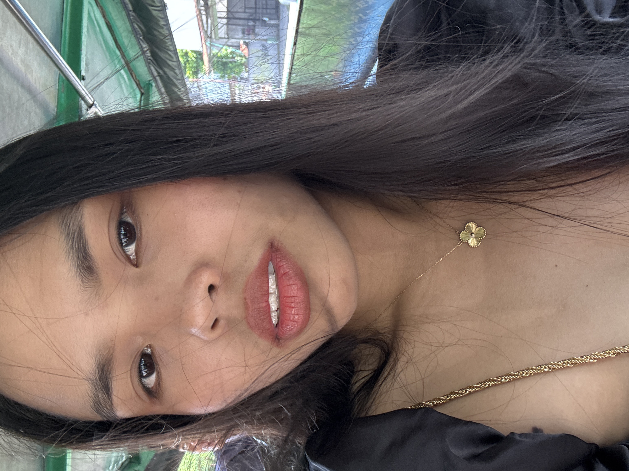 The media file features a close-up portrait of a young woman, likely of Southeast Asian descent, captured with a tilted perspective. She has long, dark brown hair that falls over her shoulders and partially across her face. Her dark eyes are directed towards the viewer, and her eyebrows are well-defined. Her lips are full, adorned with a reddish-pink color, and her upper teeth are partially visible through slightly parted lips.
She is wearing a dark-colored garment, possibly black, which appears to be off-the-shoulder. Her accessories include two gold necklaces: a delicate chain with a small, golden four-leaf clover pendant resting on her upper chest, and a thicker, braided gold chain draped lower.
The setting appears to be an outdoor or semi-outdoor location, illuminated by natural daylight. The background is softly blurred but reveals elements such as green tarp or awning structures on the upper left, suggesting a covered area like a market, a street food stall, or an outdoor cafe. Beyond these structures, indistinct shapes of foliage, buildings, and possibly fencing hint at an urban or semi-urban environment in Davao City, Philippines.
The woman's expression is neutral and engaging, suggesting she is posing directly for the photograph. There is no visible text in the image.