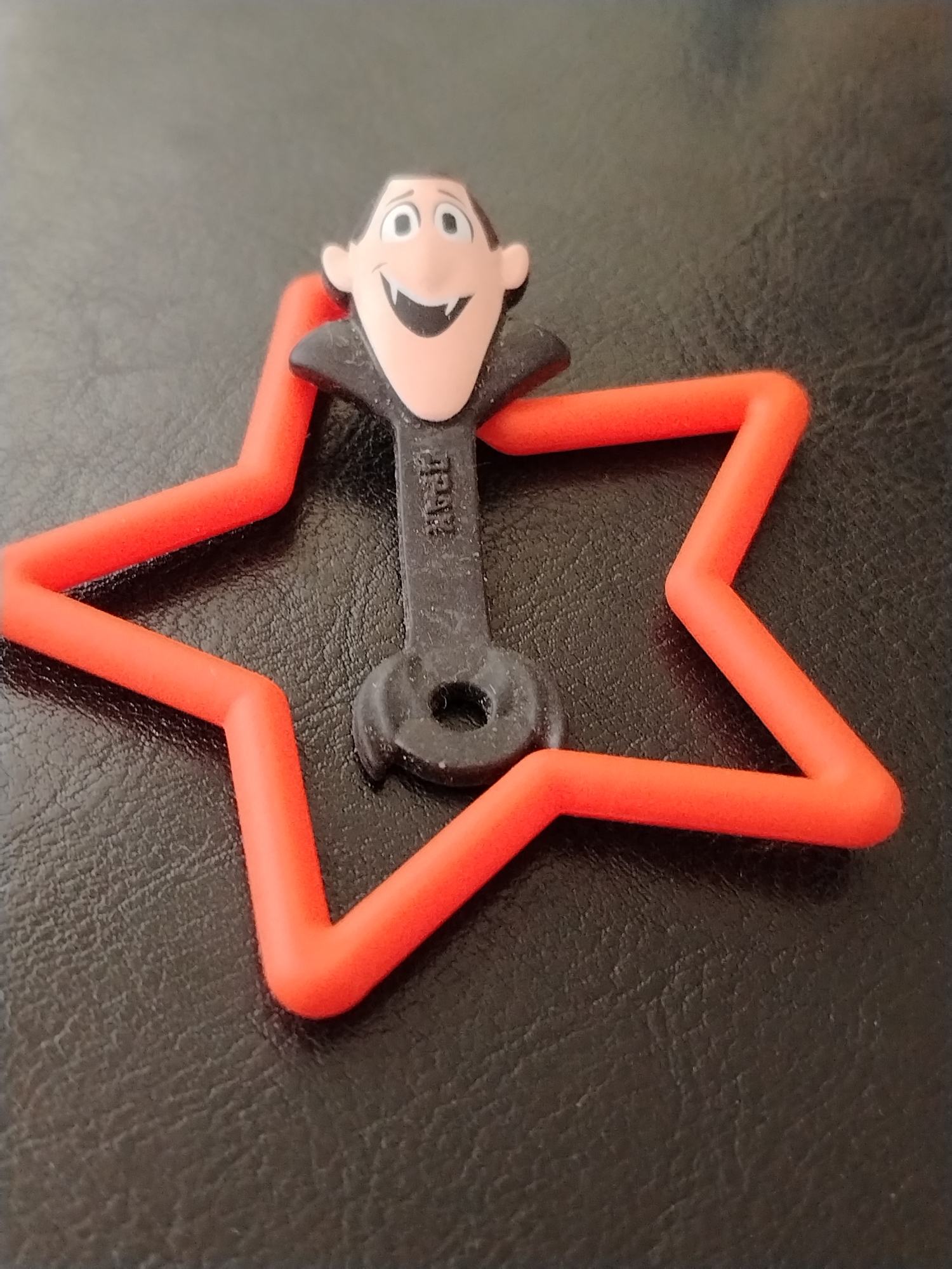 A close-up shot shows a plastic toy of Dracula, a character from the animated movie "Hotel Transylvania," positioned on a star-shaped, bright orange plastic ring. The toy, which is black and flesh-colored, has a smiling face with large eyes, prominent teeth, and slicked-back black hair. It wears a black cape. The star ring is made of a flexible, hollow plastic tube and lies flat on a dark, textured surface that appears to be faux leather. The lighting is somewhat dim, casting a soft shadow on the surface. The image does not provide any information about its location in Krasnodar, Russia.