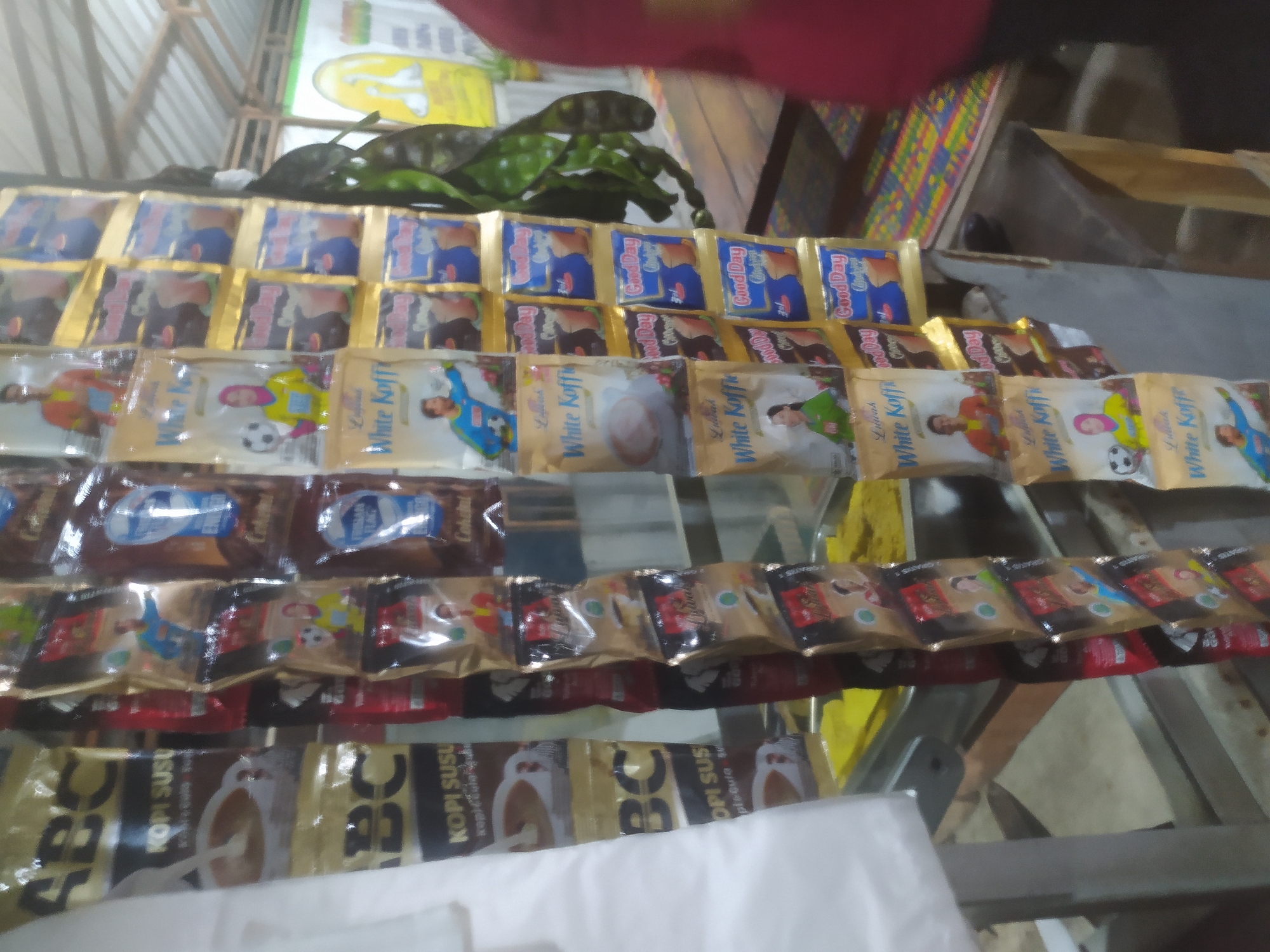 The image depicts a close-up view of a street vendor's stall in Tuntang, Indonesia. The stall is laden with numerous packets of coffee and other assorted beverages, hanging from a rack. The packets are densely packed, creating a colorful and visually busy scene. The dominant brands visible are "ABC KOPI SUSU" and "White Koffie," along with "Good Day" coffee. Some packets feature images of people, including what appear to be soccer players and women in hijabs, suggesting these are flavored coffee mixes. The background shows a rustic, open-air setting, possibly a market or roadside stall, with wooden structures and a corrugated metal roof visible. There are hints of other items for sale, such as what appears to be a bright yellow mat or tarp in the lower left, and some green produce, possibly petai beans, hanging on the right. The lighting suggests it is daytime, with a slightly overcast or shaded environment. No specific individuals are clearly identifiable or engaged in interaction, other than the implied presence of a vendor and potential customers. The overall impression is of a typical Indonesian street-side commerce scene.