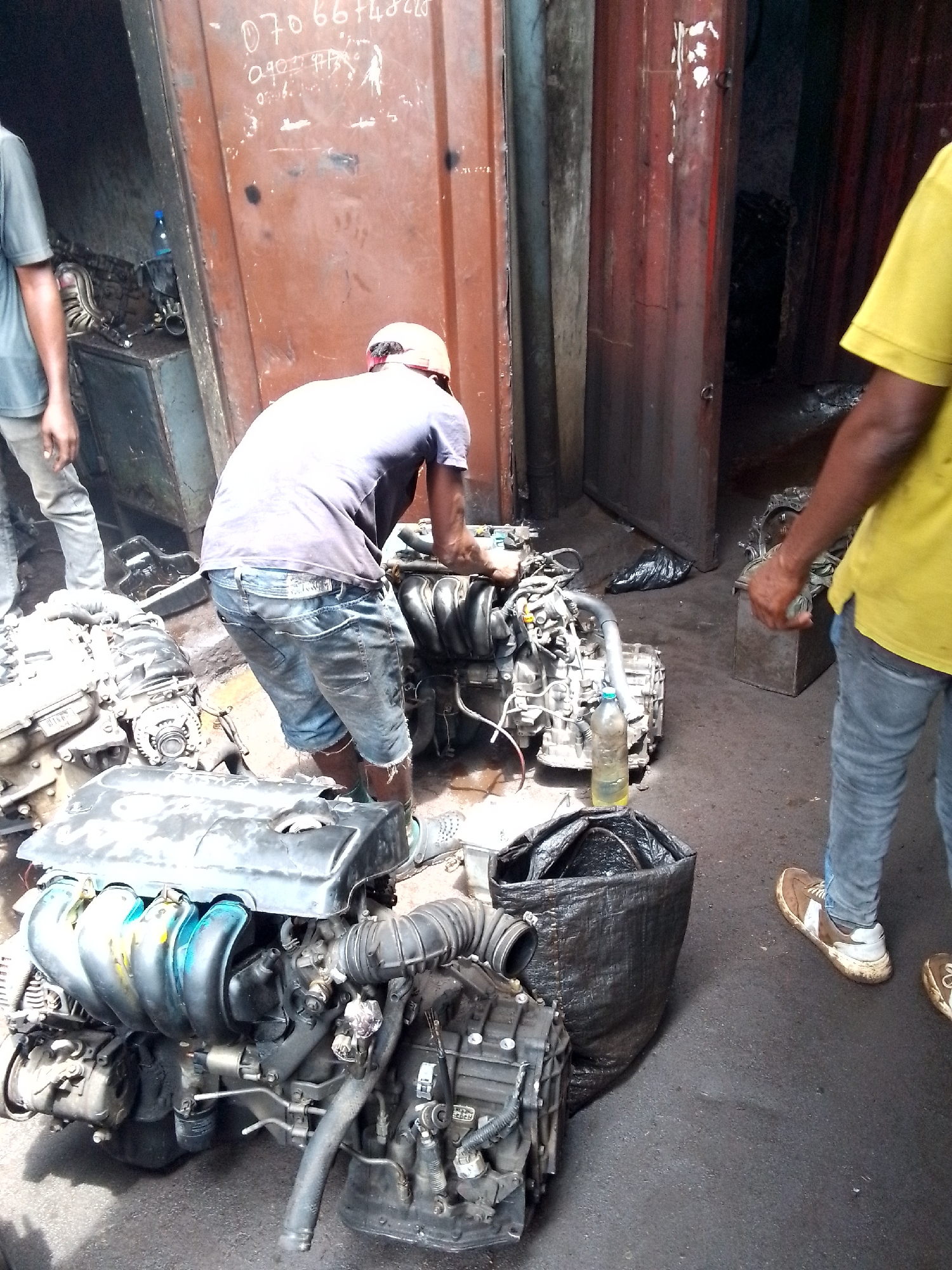 The image depicts an active outdoor or semi-outdoor auto parts market or workshop in Onitsha, Nigeria, bustling with activity around numerous car engines and automotive components. Three men are present in the scene. In the center, a man wearing a dark t-shirt, blue denim shorts, and a light-colored hard hat or cap is bending over a large car engine, seemingly working on or inspecting it. His back is to the viewer. To his left, another man in a light blue/grey t-shirt and light-colored pants stands partially visible. On the right, a third man in a yellow t-shirt and blue jeans is also partially visible, wearing brown and white sneakers.
The ground is covered with several used, greasy, and worn car engines and transmissions, indicating a business dealing in salvaged or used auto parts. A plastic bottle containing a yellowish liquid, possibly oil or fuel, is placed near one of the engines on the dark, grimy ground. Black plastic bags are also visible, one large one standing upright and another smaller one on the ground.
The background consists of tall, rusty, dark reddish-brown corrugated metal structures, likely shipping containers or large industrial doors, consistent with an informal market or industrial setting. The lighting suggests it is daytime, casting a working, functional atmosphere over the scene. Visible text on the rusty metal wall includes a phone number "070 66748220" and part of another sequence "090 77 973". The overall impression is one of industrious work in an auto parts environment.
