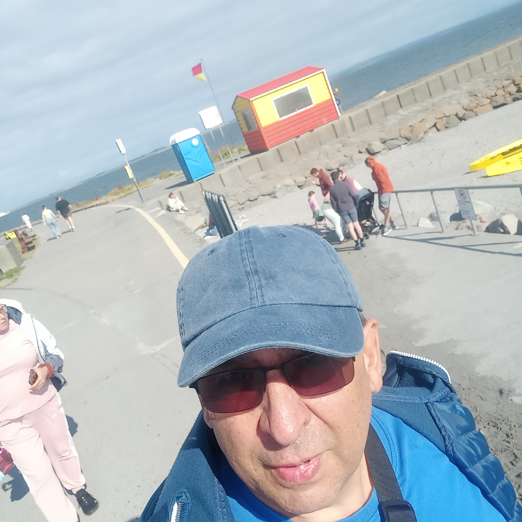 This is a first-person perspective selfie taken outdoors on a sunny day, likely in Galway, Ireland, given the presence of a brightly colored lifeguard station and a portable toilet, common at coastal areas. In the foreground, a person wearing a blue baseball cap and sunglasses is visible. They are also wearing a blue jacket over a blue t-shirt. Behind them, the scene unfolds on a paved walkway adjacent to a stony embankment leading to the sea.
Several other people are present in the mid-ground and background. To the left of the paved walkway, a person in a pink tracksuit is walking away from the camera. Further down the walkway, two figures are walking together. On the embankment, a group of people are gathered, some appearing to be children and adults.
Dominating the mid-ground on the embankment is a small, red and yellow lifeguard station, with a flag pole nearby flying a red and yellow flag. A blue portable toilet is also visible on the embankment. In the background, the calm blue sea stretches to the horizon under a lightly clouded sky. Yellow objects, possibly kayaks or buoys, are positioned on the embankment in the far right of the frame. The overall environment suggests a recreational area near the coast, possibly a promenade or a beach access point.