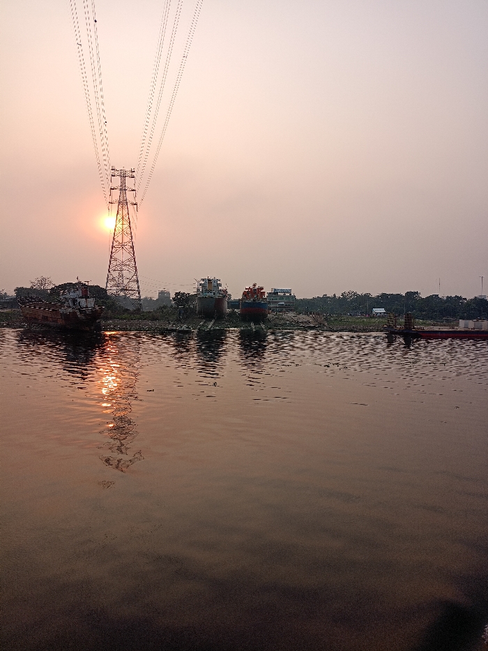 The image portrays a serene yet industrially active scene at either sunrise or sunset over a body of water, likely a river, in Zazira, Bangladesh. A towering electricity pylon stands prominently in the center-left, with its intricate structure silhouetted against the bright, low-hanging sun. Multiple power lines extend from the pylon, crisscrossing the upper part of the sky.
The sun casts a warm, orange-pink glow across the sky, which transitions from a deeper hue near the horizon to a lighter, hazy tone higher up. Its radiant reflection stretches across the water's surface, creating a shimmering path directly beneath the pylon, indicating calm conditions with gentle ripples.
Along the far bank, several boats and ships are visible. To the left, an older, possibly disused vessel rests partially on the shore, catching the sun's reflection. Further to the right, two larger ships, distinguished by their red and blue painted hulls, appear to be on land, suggesting a dry dock, shipyard, or repair facility. Trees and some structures are scattered along the distant shoreline. The water itself is still, reflecting the soft light of the sky and the industrial elements along the bank. No individuals or discernible text are visible in the photograph. The overall impression is one of an active industrial waterway bathed in the soft light of a hazy dawn or dusk.