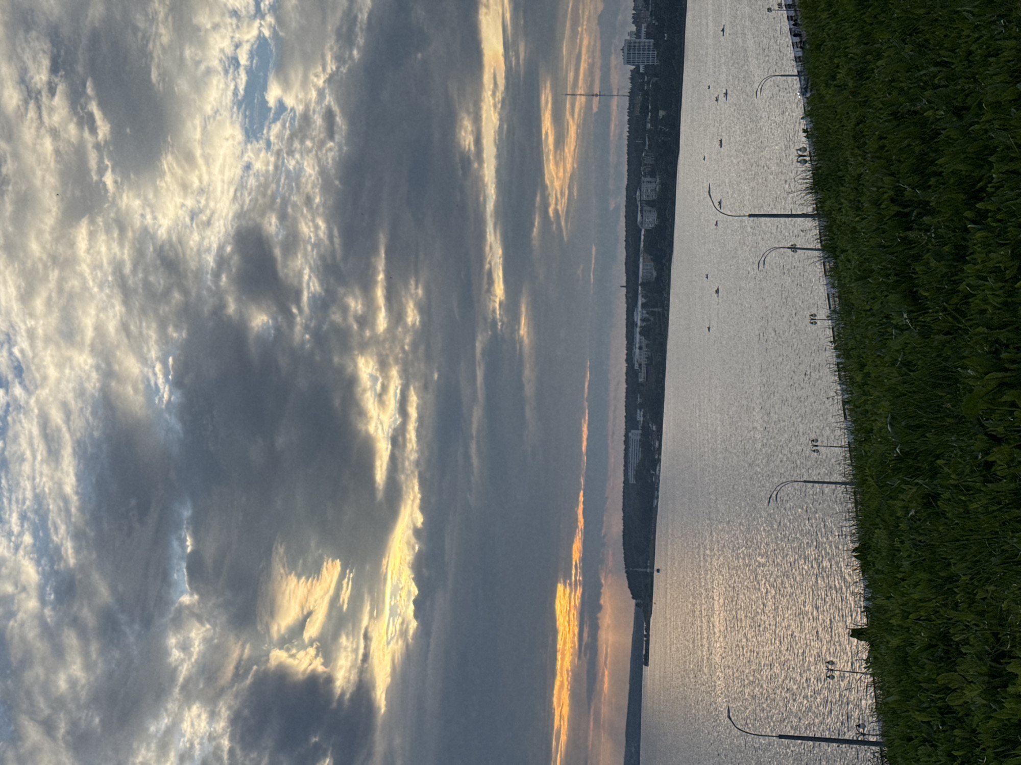 The image depicts a waterfront scene at sunset in Izhevsk, Russia. In the foreground, a lush, green, grassy slope leads down to a wide body of water. Several lampposts are visible along the embankment separating the grass from the water. On the water's surface, multiple small boats, possibly kayaks or canoes, are scattered. Across the water, a city skyline is visible, characterized by buildings of varying sizes and architectural styles, including a tall transmission tower. The sky is dramatic, filled with heavy, gray clouds interspersed with streaks of bright, golden sunlight, suggesting the sun is setting or has recently set. The overall impression is one of natural beauty combined with an urban landscape.