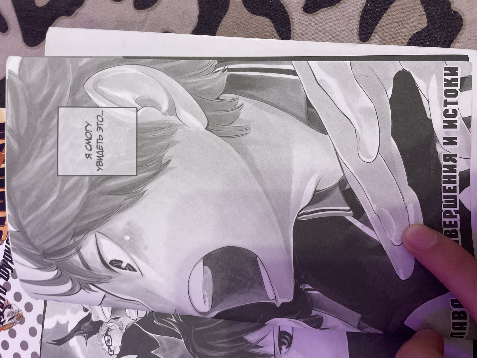 The image shows a close-up of a manga panel held by a person's finger. The manga is black and white and features a close-up of an anime-style character's face, looking upwards with an open mouth and large eyes. Text is visible on the left side of the manga panel in Russian, reading "ВЕРШЕНА И ИСТОКИ" and "ЛАБО". A small rectangular box with Russian text is visible on the right side of the panel, reading "Я СМОТРЮ УВИДЕТЬ ЭТО...". The background of the image is a camouflage pattern fabric. There is no indication of the city of Irinovka, Russia, or any specific environmental context beyond what is depicted on the manga and the immediate foreground.