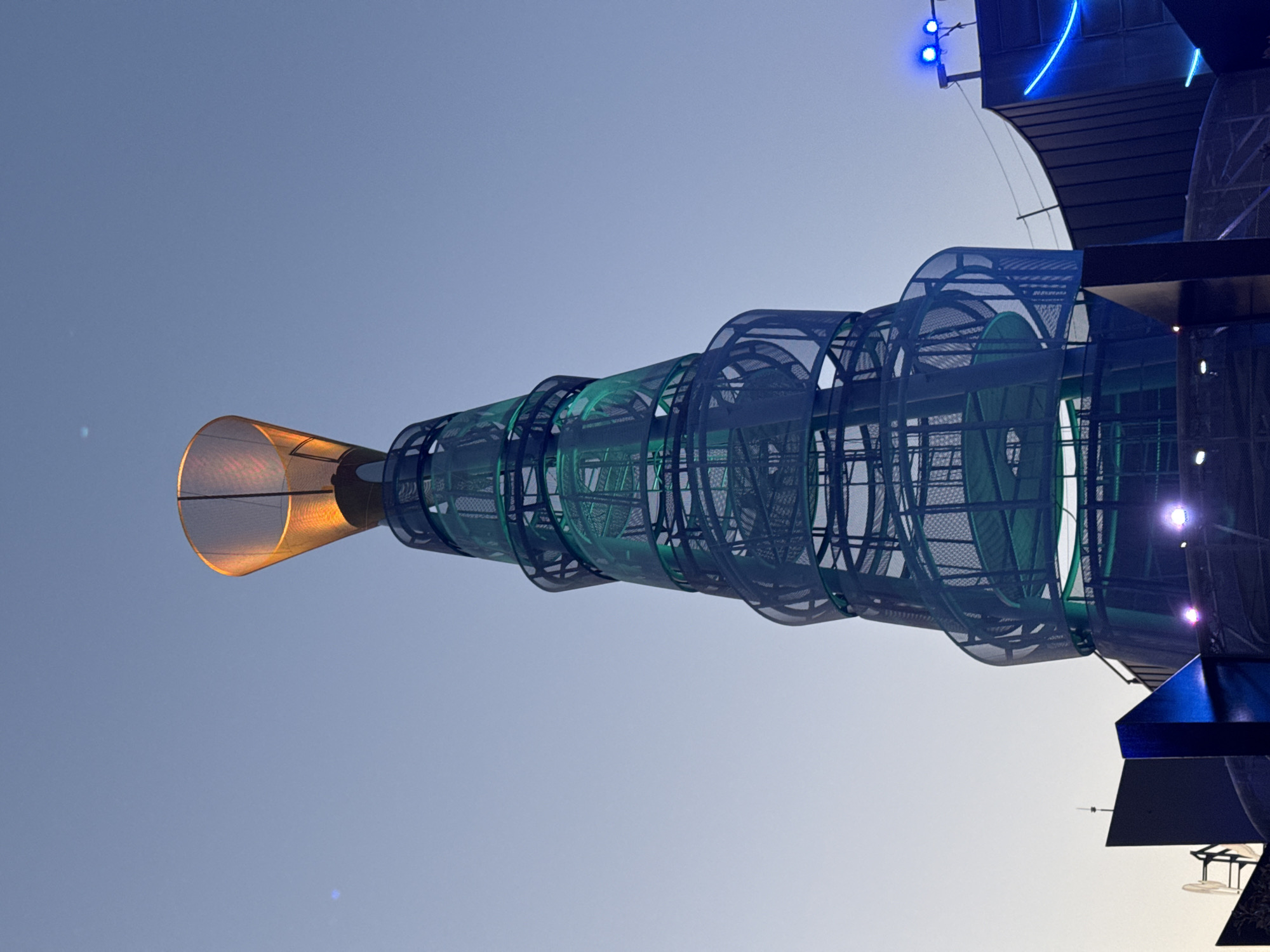 An outdoor, low-angle, full shot captures a towering, spiraling structure against a clear, pale blue sky. The structure, composed of dark metal mesh and green illuminated platforms, resembles a twisted funnel or a giant corkscrew. At its apex, a cone-shaped element glows with a warm, orange light. The base of the structure is surrounded by dark, angular architectural elements and illuminated with blue and pink lights. The scene appears to be an amusement park or an outdoor event venue. The description is consistent with images of "The Wave" at the Heron City entertainment complex in Las Rozas de Madrid, Spain.