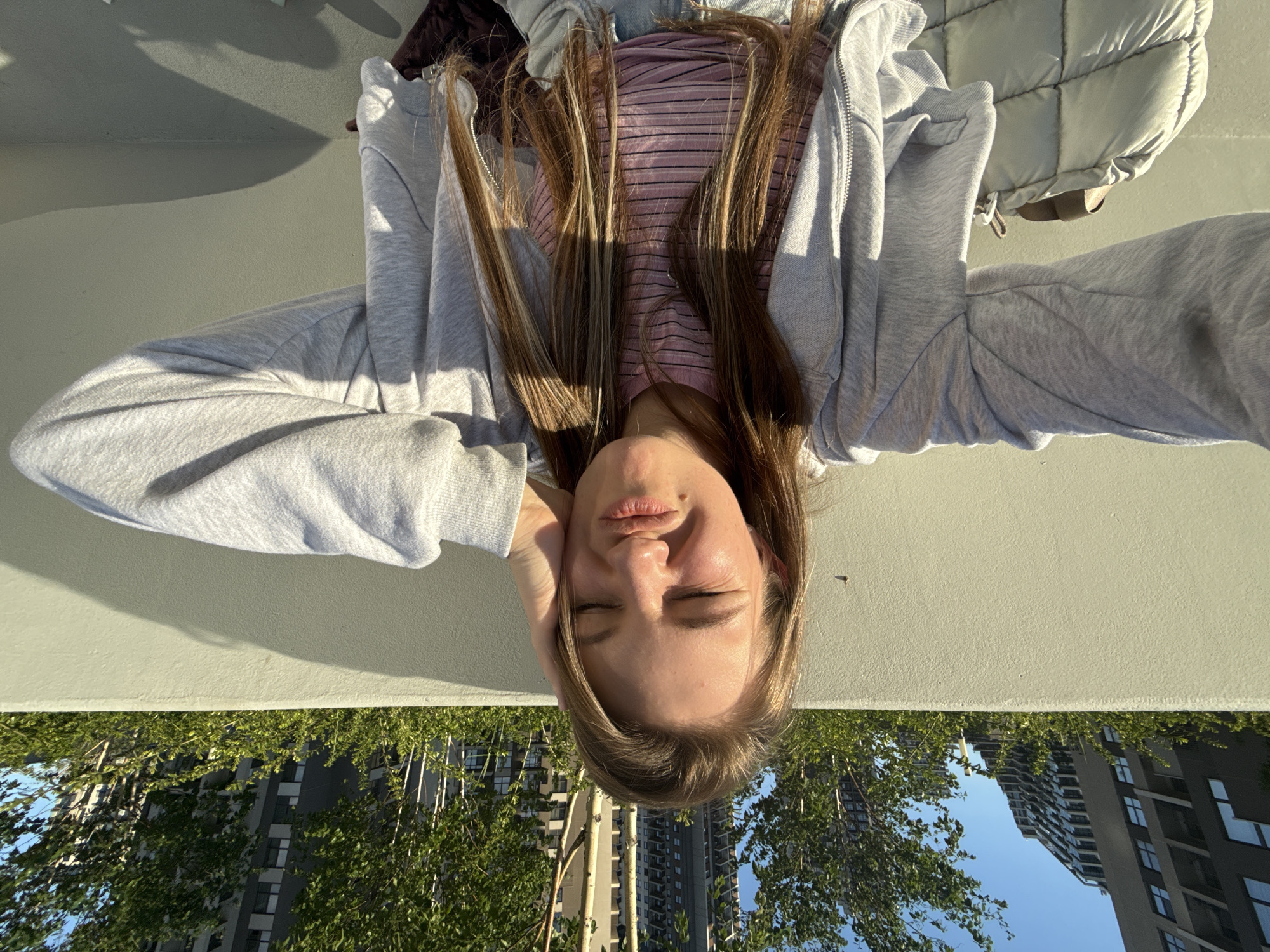 The image is an inverted selfie of a young woman lying outdoors on a sunny day.
The subject is a young woman with long brown hair, featuring blonde highlights, spread around her head. Her eyes are closed, and her lips are slightly parted, conveying a serene or pensive expression. She is wearing a light grey zipped hoodie over a pink striped top. A light-colored, possibly white or light grey, puffer vest or jacket is visible behind her head and near her right shoulder (left side of the image as displayed). Her hands are gently resting on either side of her head.
When the image is viewed in its intended orientation (rotated 180 degrees), the woman is looking upwards. Above her are several tall, modern buildings with visible windows, interspersed with vibrant green deciduous trees against a clear blue sky. The lower part of the image shows a light-colored, textured wall or ground surface, likely concrete, upon which she is resting.
The scene is brightly lit by direct sunlight, casting distinct shadows on her face, hair, and clothing, as well as on the resting surface. This indicates a clear, sunny day, likely mid-day or late afternoon. The setting is an urban or suburban area, consistent with Belgrade, Serbia. No visible text is present in the image.