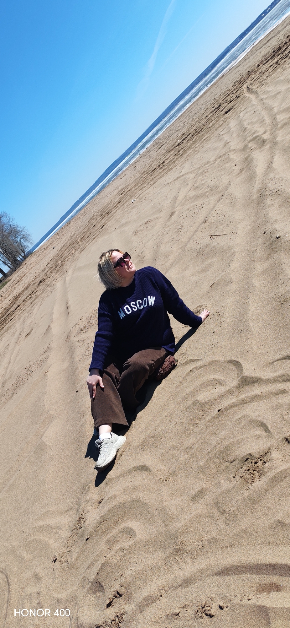 A woman with blond hair and sunglasses is sitting on a sandy dune. She is wearing a dark blue sweater with the word "MOSCOW" in white letters, brown pants, and white sneakers. The dune appears to be near a beach, with the ocean and a clear blue sky visible in the background. The scene is brightly lit. The camera that took the photo is an Honor 400. The scene is located in Noviy, Russia.