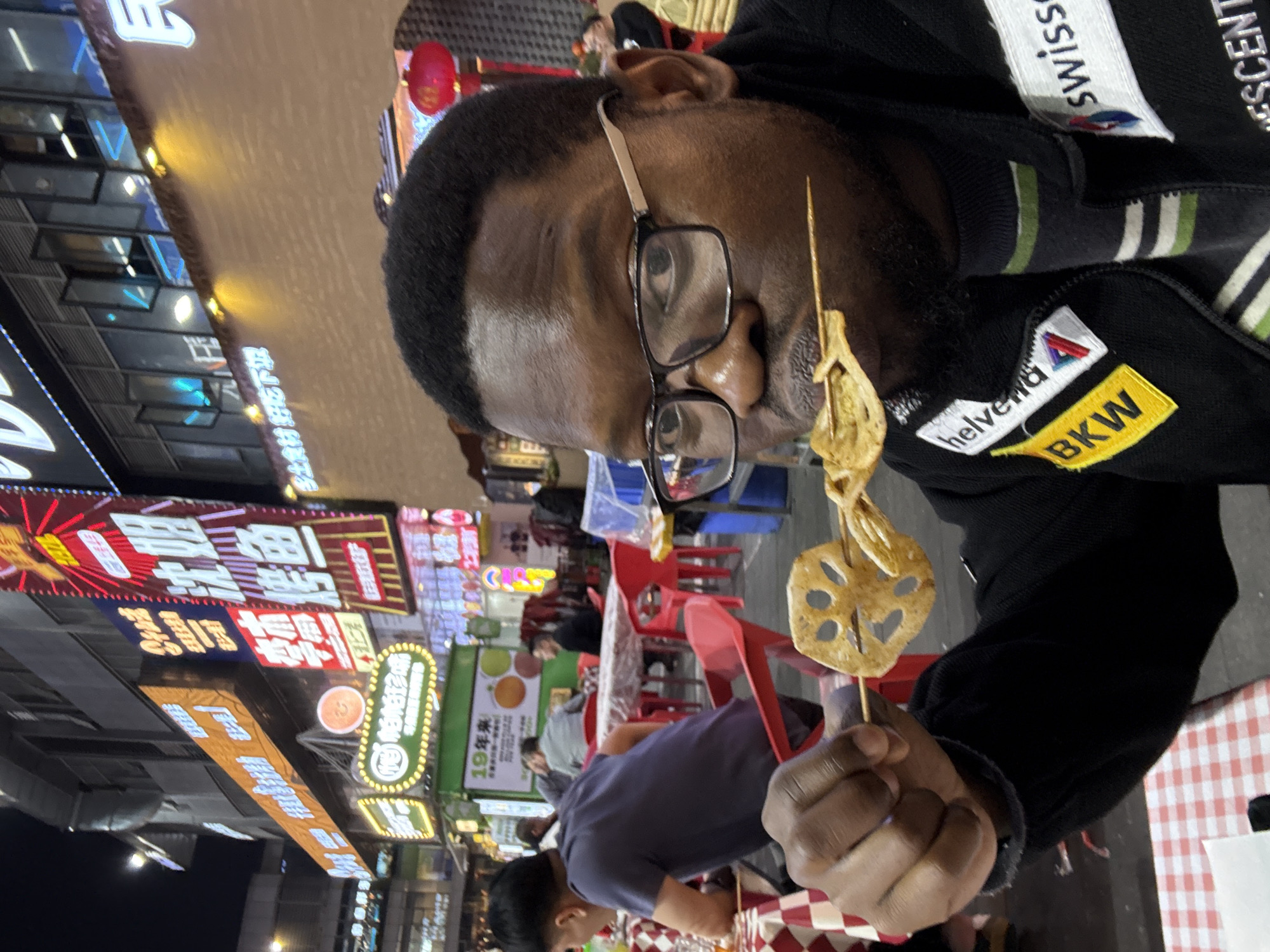 The image depicts a nighttime outdoor dining scene in Liangjiang, China. In the foreground, a dark-skinned male subject with short, dark hair and a beard, wearing black-framed eyeglasses with silver temples, looks directly towards the viewer. He is dressed in a black zippered jacket adorned with white "helvetia" and "swisss" patches, and a yellow "BKW" patch, over a striped shirt. The subject holds a wooden skewer with two pieces of food, identified as sliced lotus root and another cooked flat item, positioned in front of his mouth.
The immediate foreground includes a table covered with a red and white checkered tablecloth. The background features a bustling urban street with multiple multi-story buildings. These buildings are heavily adorned with numerous illuminated neon and LED signs displaying Chinese characters, including a prominent vertical sign that reads "沈姐 烤鱼" (Sister Shen Roasted Fish). Other signs visible include "19年来" (19 years). Several individuals are seated at outdoor tables equipped with red plastic chairs and similar checkered tablecloths. One man in a dark blue shirt is seated with his back to the camera at a table to the left. A red Chinese lantern hangs in the upper right background, contributing to the ambient lighting from the various signs and street fixtures.