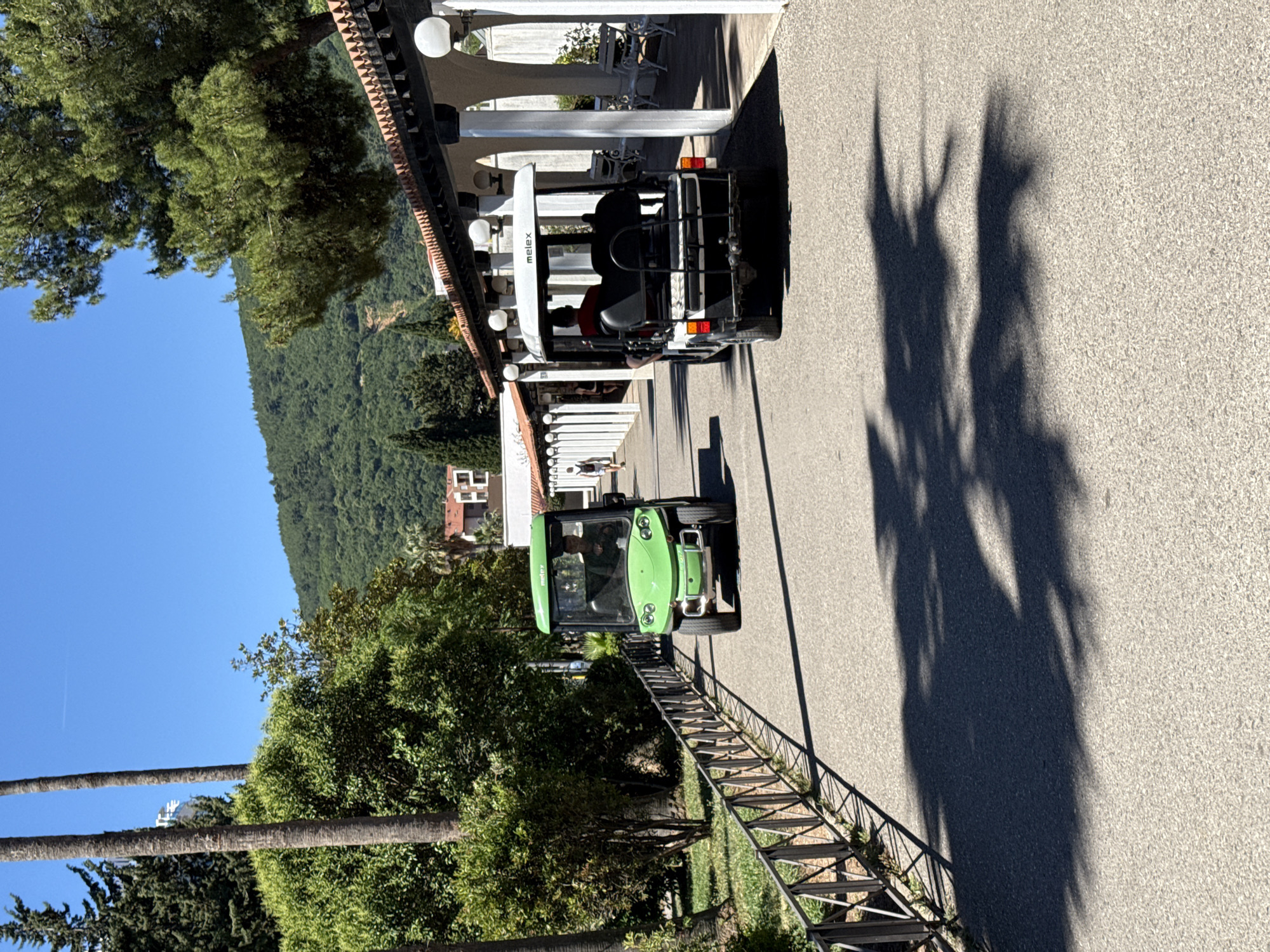 The image shows a street in Budva, Montenegro with two vehicles: a white Melex utility vehicle and a green utility vehicle. The Melex is parked in front of a building with a white facade and an arched colonnade. The green vehicle is further down the road, closer to a metal railing separating the road from a grassy area with trees. Shadows of trees are cast on the asphalt. In the background, a hill covered in trees is visible under a blue sky.