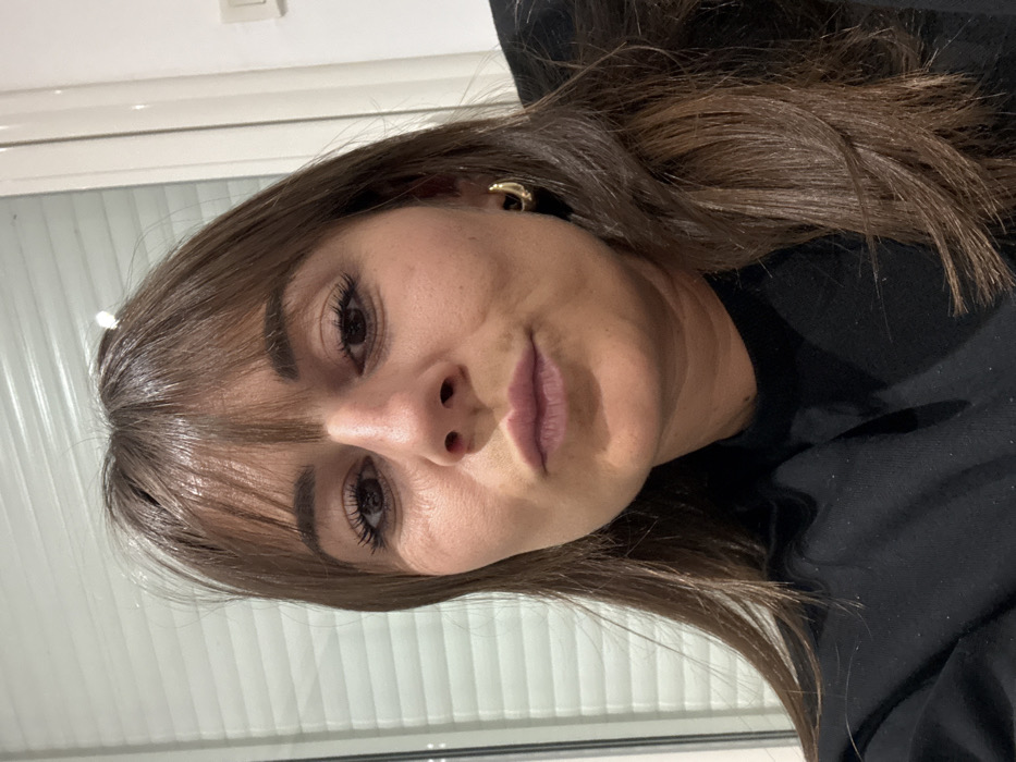 A woman with light skin and brown hair poses for a portrait. She has dark eyes with long eyelashes and a straight nose. Her lips are a pale pink and her hair is styled with bangs. She wears a gold earring. A window with blinds is visible in the background. She is wearing a black shirt.