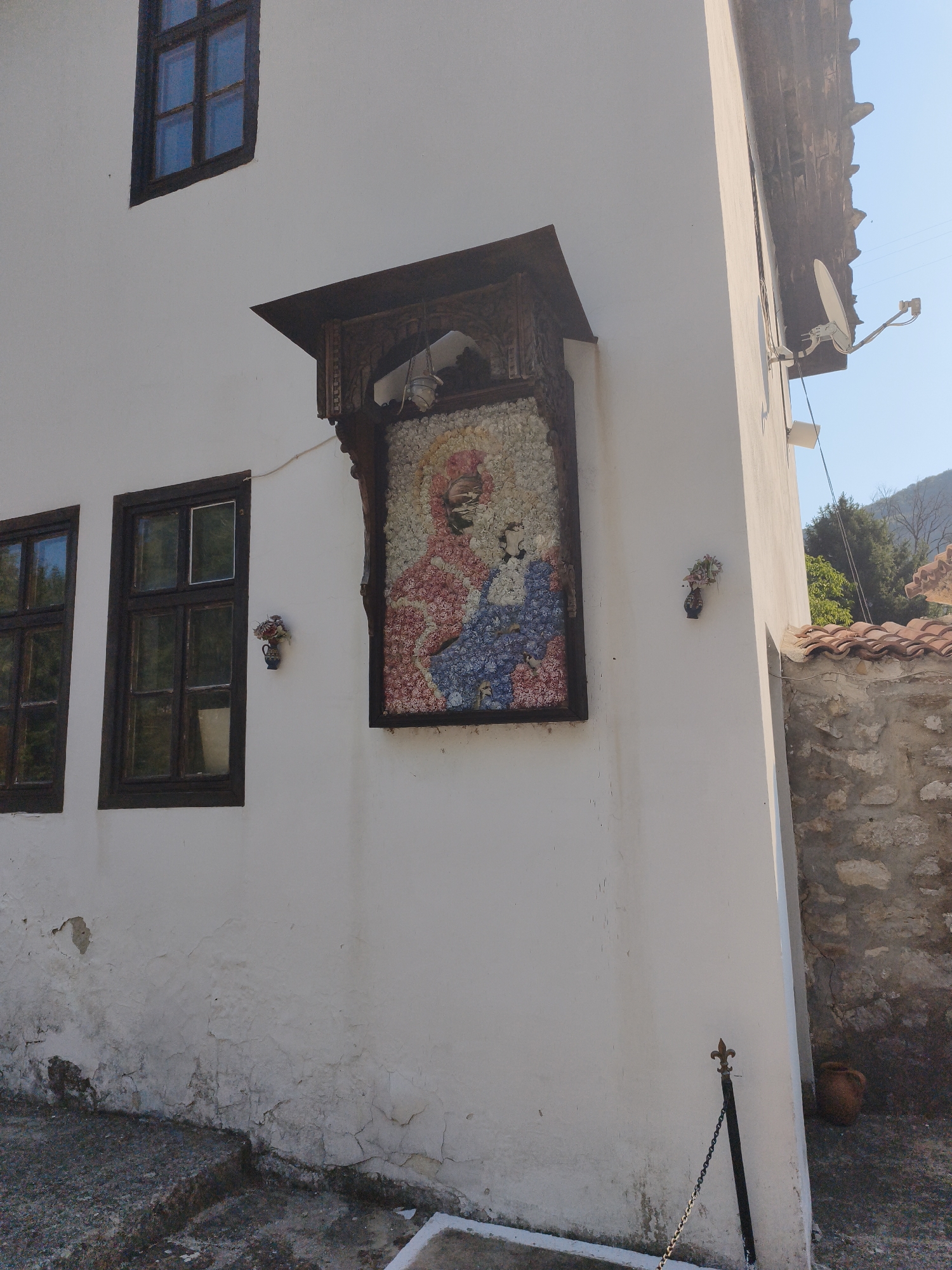 The image depicts the exterior of a white building with a dark-tiled roof and a stone wall on the right side. A decorative mosaic image of what appears to be a religious figure, possibly the Virgin Mary and child, is affixed to the white wall. This mosaic is framed by an ornate wooden structure. There are two windows with dark wooden frames on the left side of the building. Small decorative vases with floral arrangements are attached to the wall near each window. A satellite dish is mounted on the upper right section of the building. The ground in front of the building is a rough, textured surface with some steps. A metal post with a chain attached is visible near the bottom right corner of the building. The overall scene suggests an architectural detail or facade in Lyutibrod, Bulgaria.