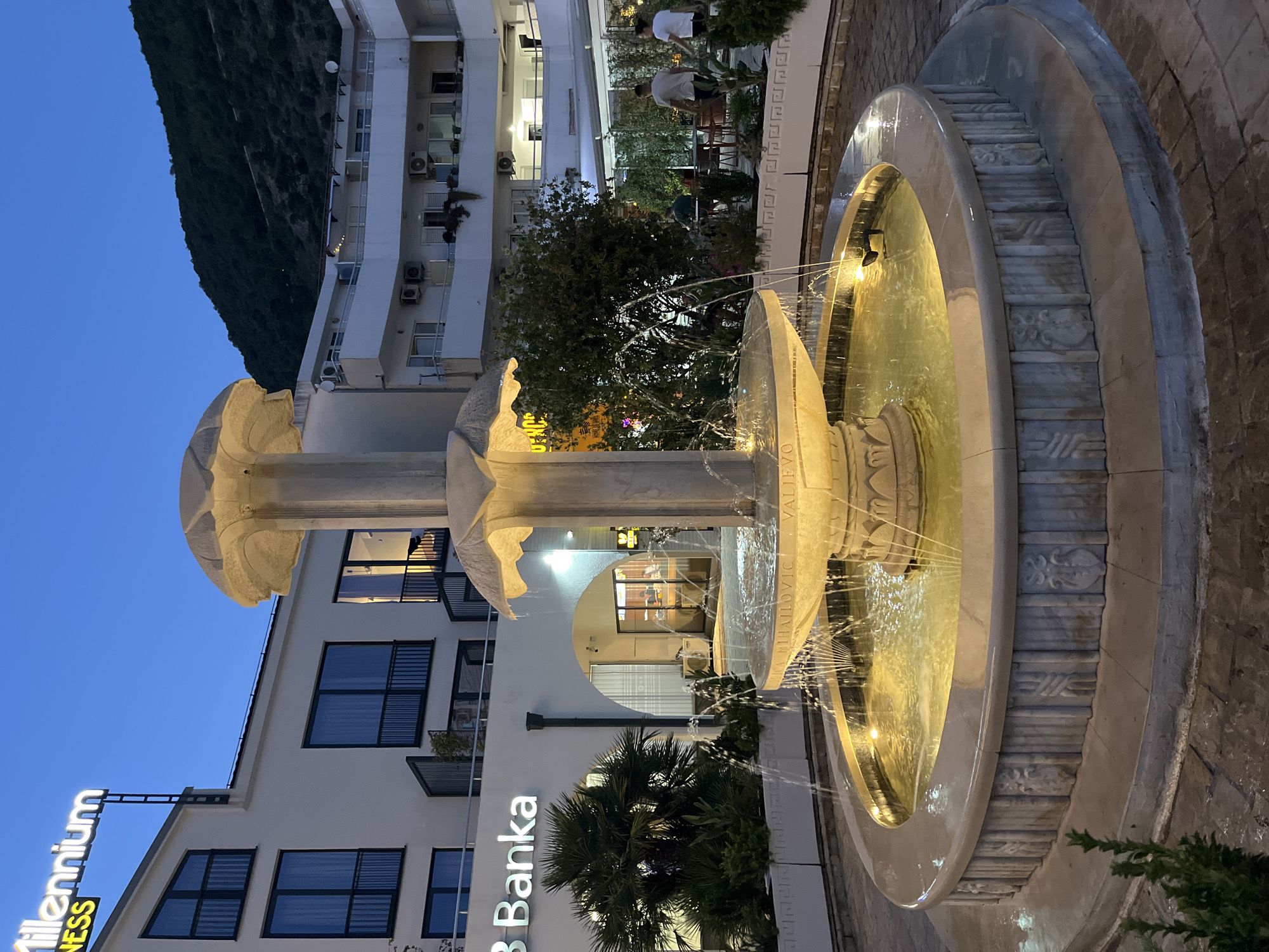 The image shows an outdoor fountain in the city of Budva, Montenegro. The fountain is multi-tiered and made of stone, with water cascading down its levels. It is illuminated by warm yellow lights, creating a soft glow in the twilight. Surrounding the fountain is a paved area with decorative stone elements on the fountain's base. In the background, there are several buildings. To the left, a white building with a neon sign that reads "Millennium Fitness" is visible. Below the sign, the text "B Banka" is also illuminated. The buildings have multiple stories with balconies and windows. To the right, another building with balconies and air conditioning units is visible, and beyond that, a green hillside rises. In the middle ground, there is an outdoor seating area with tables and chairs, where a few people are seated and two individuals are walking. The overall scene suggests an evening ambiance in a public space.