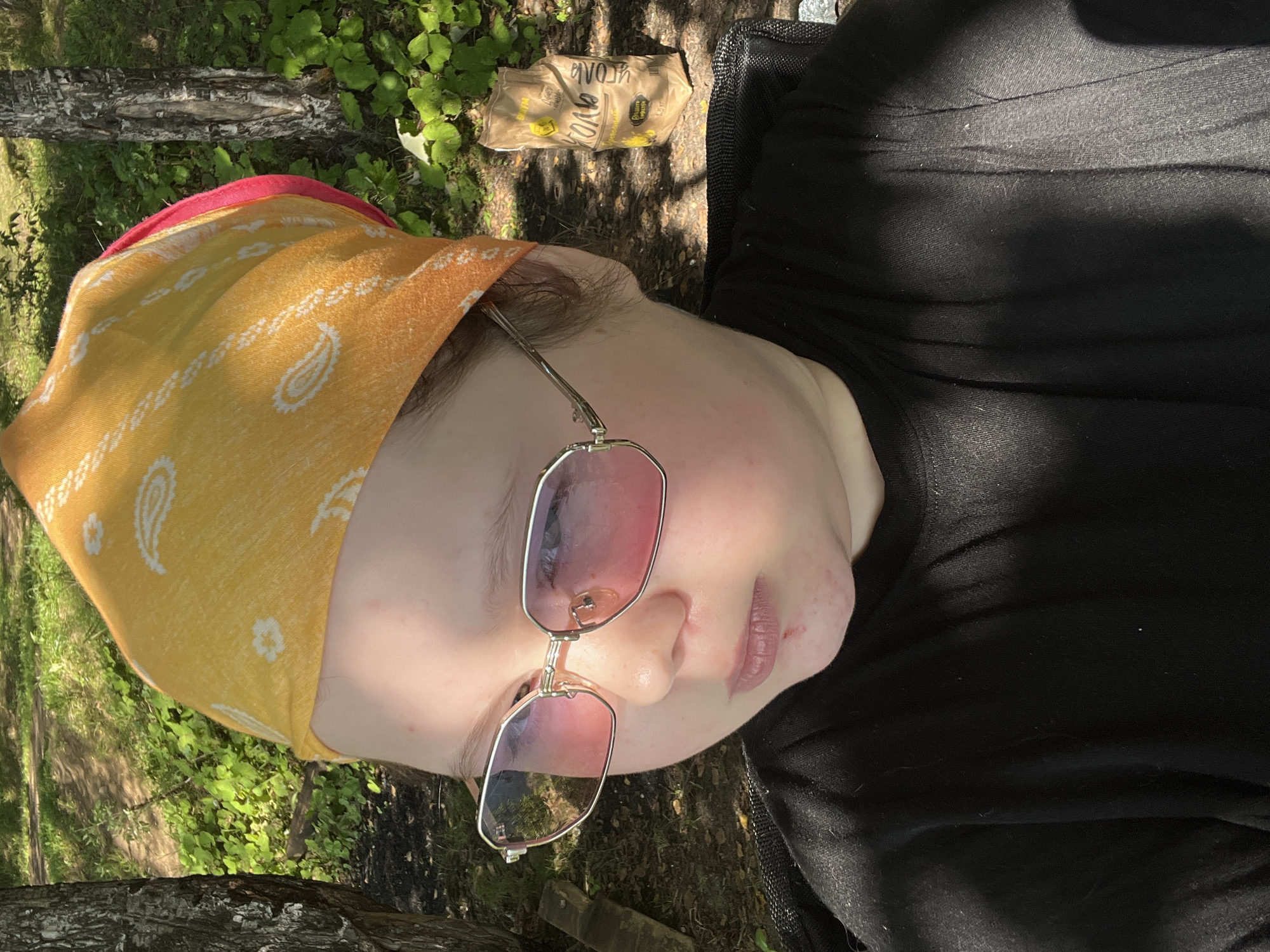 This media file depicts a person lying down outdoors, with a close-up view of their face and upper body. The subject is wearing a yellow bandana with a paisley pattern and pink-tinted sunglasses. A black shirt is visible, along with a portion of what appears to be a dark bag or backpack.

The background consists of lush green foliage and trees, suggesting a natural, wooded setting. Sunlight filters through the leaves, casting shadows and creating dappled light on the subject's face and the ground. This indicates it is daytime, likely sunny.

On the ground near the subject, a crumpled brown paper bag is visible. The bag has some text printed on it, but it is not clearly legible in the image. The overall impression is one of relaxation in a natural environment. The image appears to be taken from a slightly elevated angle, looking down at the subject. The person's face shows a neutral or relaxed expression.

Location context of Romashka, Russia, suggests a rural or natural area. The presence of trees and green plants aligns with this. No specific activity is clearly depicted, beyond the subject resting. The time of day is indicated by the sunlight and shadows. No strong emotions are conveyed, though a sense of calm might be inferred from the pose and setting.