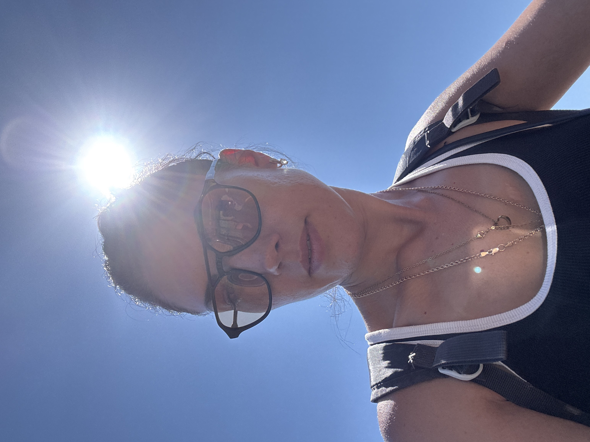 A woman is photographed from a low angle, with the sky and sun in the background. She is wearing black-framed glasses, a black tank top with white trim, and multiple gold necklaces. Straps from her top cross over her shoulders. The sun is positioned to the left of her head, causing a bright flare. The background is a clear blue sky. The location is most likely the Hof HaCarmel Regional Council, Israel.