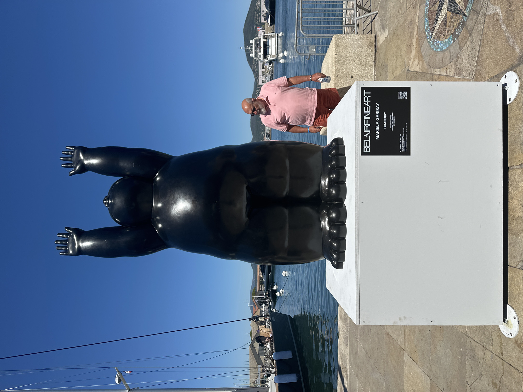 An outdoor, eye-level, medium shot captures a large, black, glossy bronze sculpture of a plump, humanoid figure with its arms raised high and palms facing upwards. The sculpture stands on a white rectangular pedestal in a marina. A man with a shaved head, wearing a pink polo shirt and orange shorts, stands to the right of the sculpture, holding a small white box. In the background, several sailboats and yachts are docked in the blue water under a clear blue sky, with a city skyline and green hills in the distance. The scene appears to be at the Port of Saint-Tropez, France.