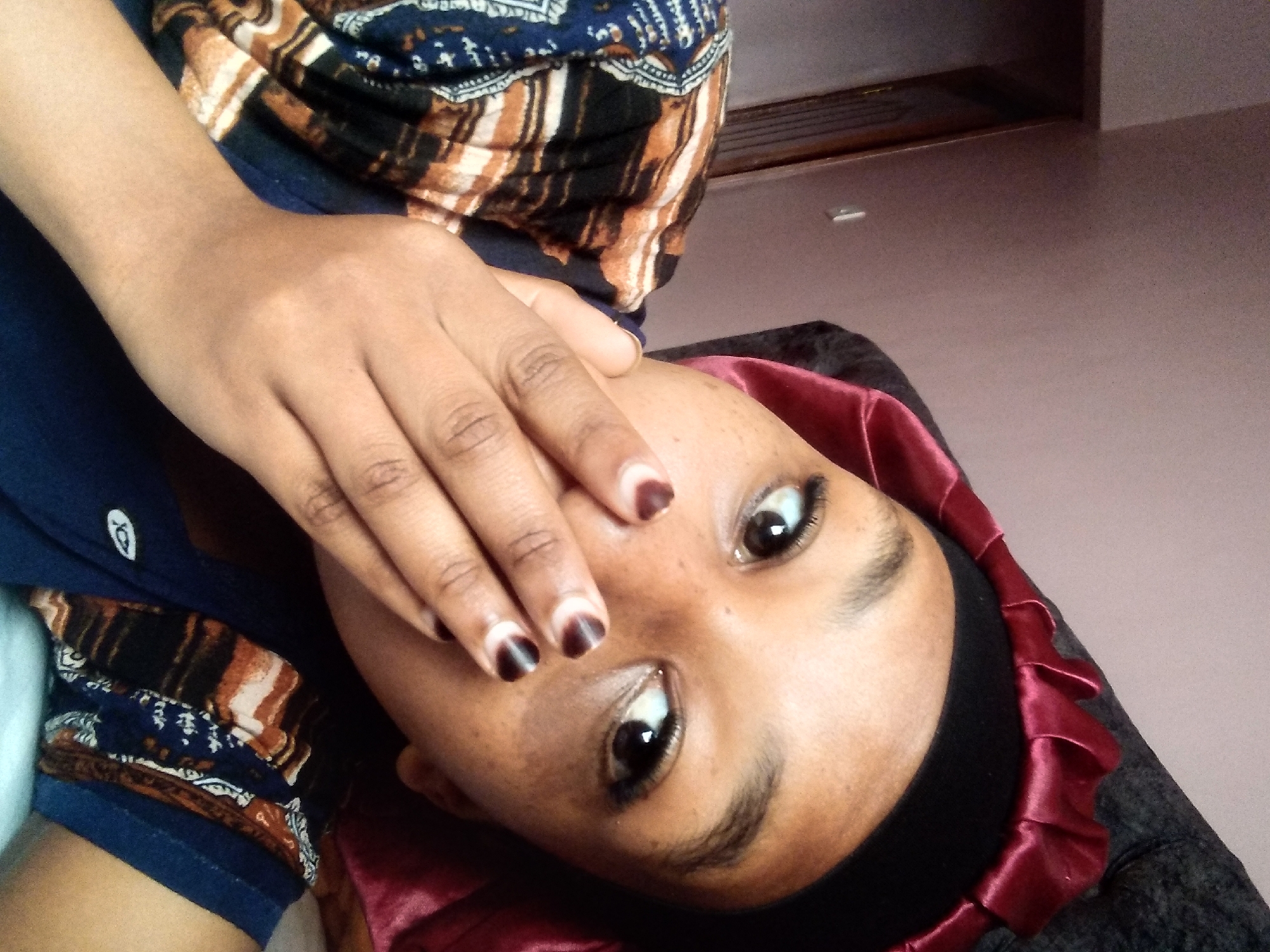 A young Black person, possibly a female, is shown in a close-up, waist-up shot from a slightly elevated angle. They are wearing a black headscarf with a burgundy satin trim that is ruffled. A patterned garment with blue, white, black, and orange sections is visible over their shoulders. Their right hand is covering their mouth, and they are wearing dark nail polish with a white crescent shape at the base of each nail. The person's eyes are open and looking towards the camera. The background appears to be an indoor setting with a textured, light-colored wall and a dark surface, possibly a piece of furniture or bedding, behind them. There is no indication of the city of Nyeri, Kenya, in the image itself.
