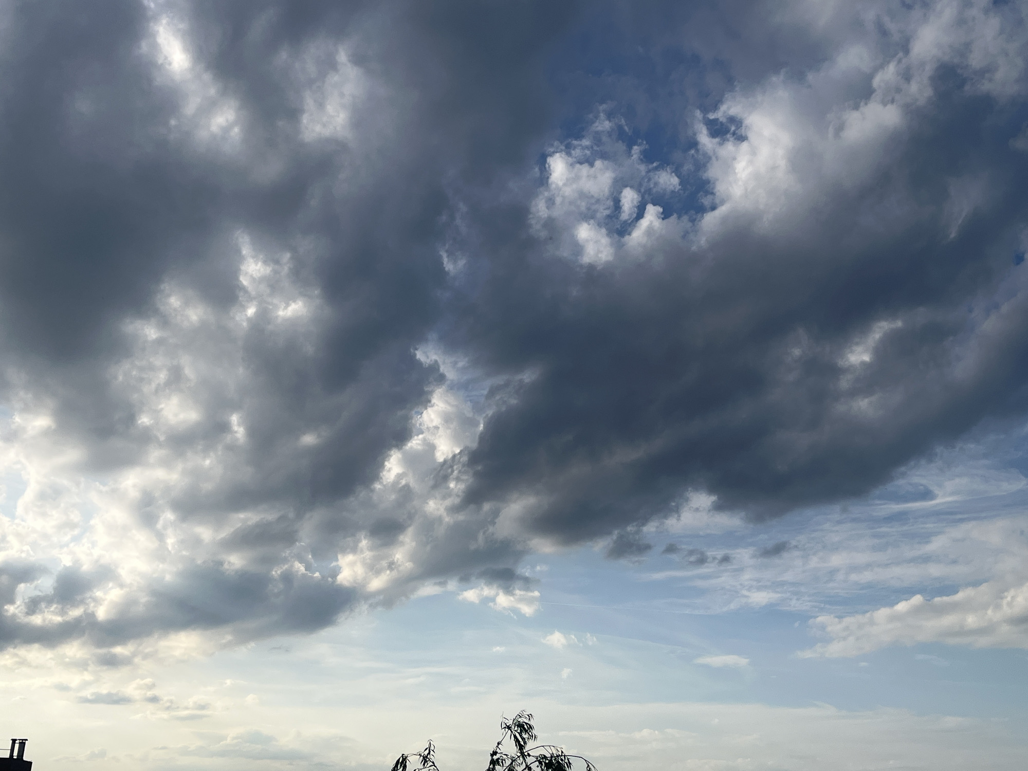 The image shows a sky filled with various types of clouds, ranging from dark, heavy storm clouds to lighter, wispy clouds. There's a patch of blue sky visible amidst the clouds. In the bottom of the image, there are silhouettes of trees and a building. The location is in the city of Unknown, Unknown.