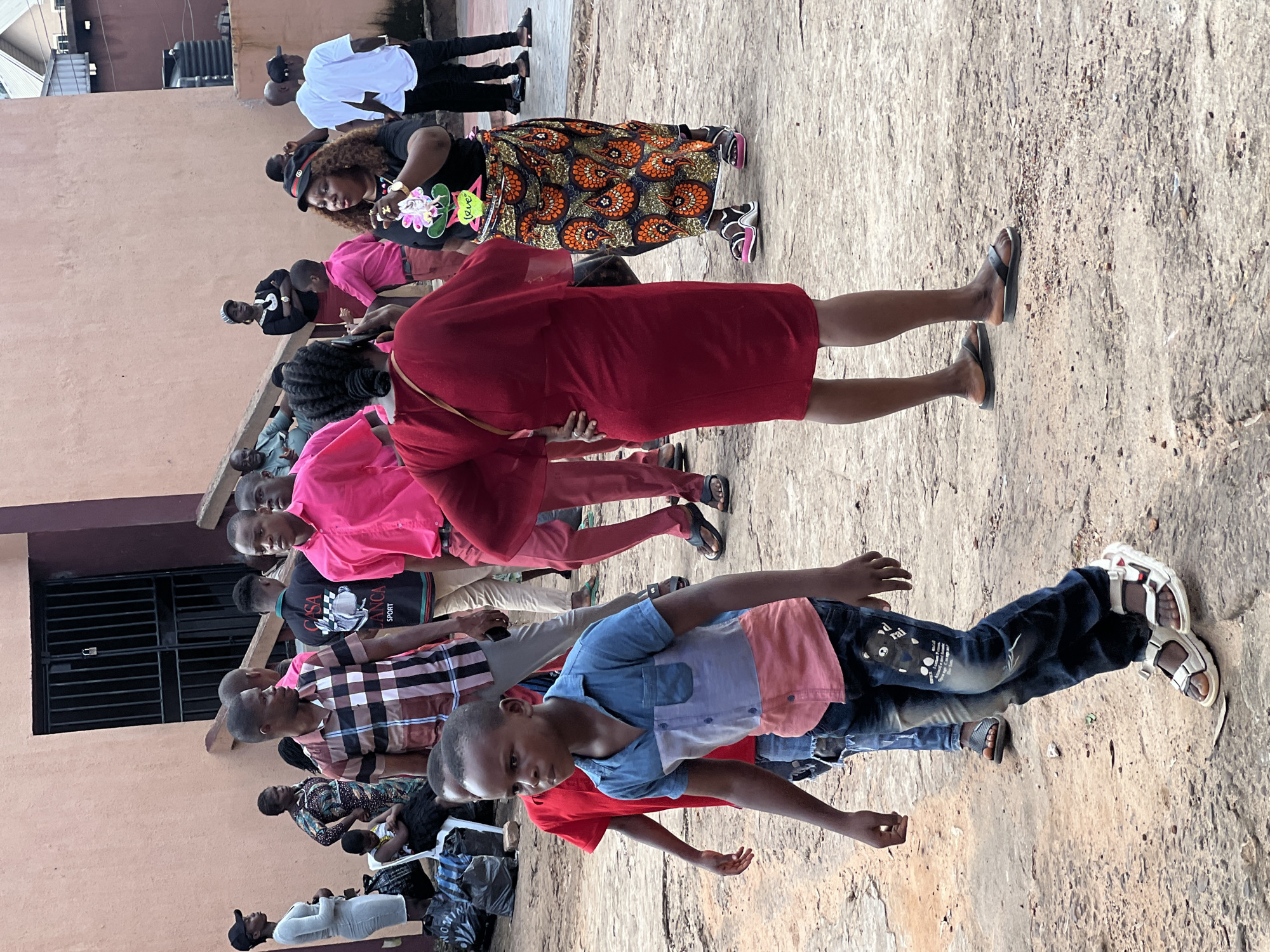 In Nnewi, Nigeria, a group of people are gathered on a dusty, outdoor surface in front of a peach-colored building with a dark metal-barred door. The foreground features a young boy in a blue and pink color-blocked shirt and dark jeans walking towards the left, his gaze directed forward. To his right, another boy in a pink shirt and dark jeans walks in the same direction.
Further back, a woman in a vibrant red dress with flowing sleeves stands with her hands on her hips, facing away from the camera. Next to her, another woman in a black t-shirt with a floral design and a patterned skirt is speaking, gesturing with her right hand. A black hat with a brim is perched on her head. Behind them, several men and women are visible, some in brightly colored shirts, others in white. Bags are scattered on the ground to the left, near a white plastic chair where a person is seated. The environment suggests a public gathering or event.