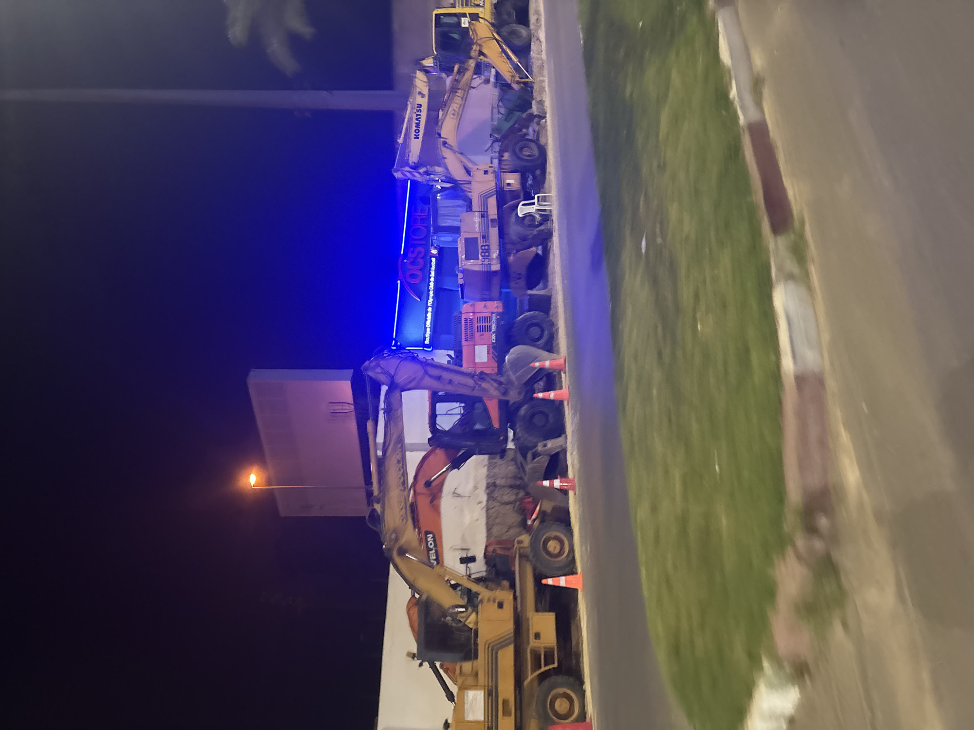 At night in an unknown city, several heavy construction vehicles, including excavators and a backhoe loader, are parked in front of a building with blue lighting. One of the excavators has the brand name "KOMATSU" visible. A sign above the building reads "OCSTORE" and "Boutique Officielle de l'Olympic Club de Sari Footbal". Orange traffic cones are placed around the construction vehicles. The ground is paved with asphalt, and a grassy area with a curb is visible in the foreground. A single white plastic chair is placed near one of the vehicles. A street lamp with a warm yellow glow is also present.