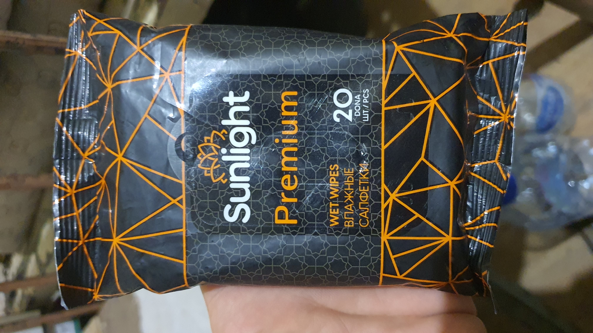 The image shows a close-up of a black and orange package of Sunlight Premium wet wipes. The package has a geometric pattern of intersecting orange lines on a black background. The Sunlight logo, a stylized lotus flower in orange, is visible above the word "Sunlight" in white, sans-serif font. Below "Sunlight" is the word "Premium" in large, orange, serif font. Further down, "WET WIPES" is written in white, and below that, in a Cyrillic script, are the words "ВЛАЖНЫЕ" and "САЛФЕТКИ". To the right of the text, the number "20" is displayed in large white digits, with "DONA" and "ШТ/PCS" written below it in smaller text. The package is being held by a hand, partially visible on the left side of the frame. In the background, out of focus, are several plastic water bottles with blue caps. The image appears to be taken indoors, possibly in a shop or a home setting, with some blurred wooden structures visible in the upper background. There is no indication of the city of Karankul, Uzbekistan, in the image.