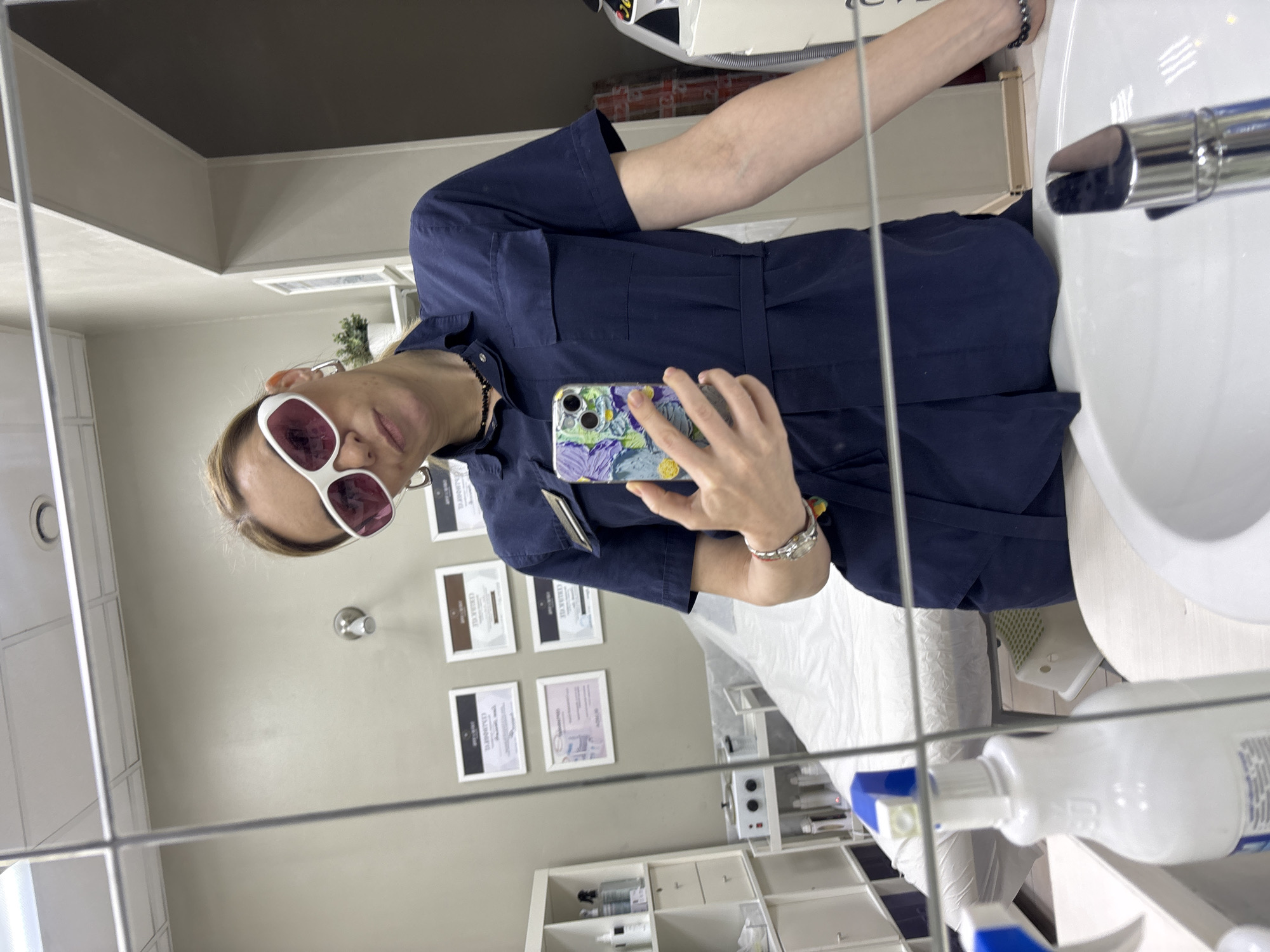 A person is reflected in a mirror within what appears to be a commercial or healthcare setting, potentially located in Vsevolozhsk, Russia. The person is wearing sunglasses with white frames and rose-tinted lenses and a dark blue uniform-style garment. They are holding a phone with a decorative blue and yellow case, presumably taking the photograph. The background features wall-mounted certificates or documents, a shelf with various bottles, and what looks like a medical or cosmetic bed covered with white material. A sink and faucet are visible in the foreground of the reflection. The scene suggests a clinic, salon, or medical office.