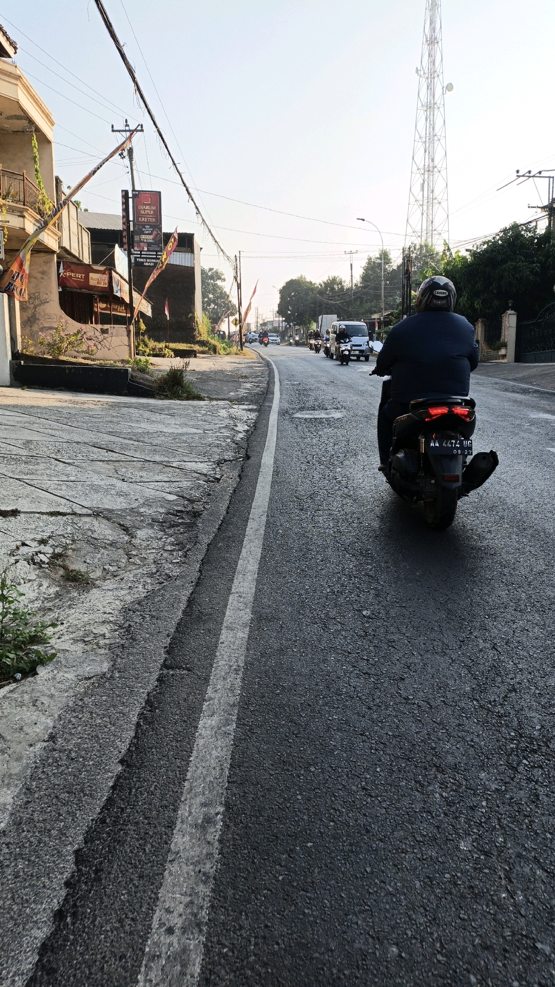 In Kuwasen, Indonesia, a lone motorcyclist with a helmet is captured from behind, riding a dark-colored scooter down an asphalt road. The scooter has a license plate that reads "AA 4674 UC" and a visible expiration date of "09-27". The road is marked with a white double line and is flanked by a cracked concrete embankment on the left, where some sparse vegetation grows. In the background, other vehicles, including a white van and several motorcycles, are visible traveling in the same direction. A tall transmission tower stands prominently in the distance, silhouetted against a hazy sky. The scene appears to be in a semi-urban or rural setting, with buildings and trees lining the roadside. The lighting suggests it is daytime, possibly late morning or early afternoon, given the angle of the sun and the shadows.