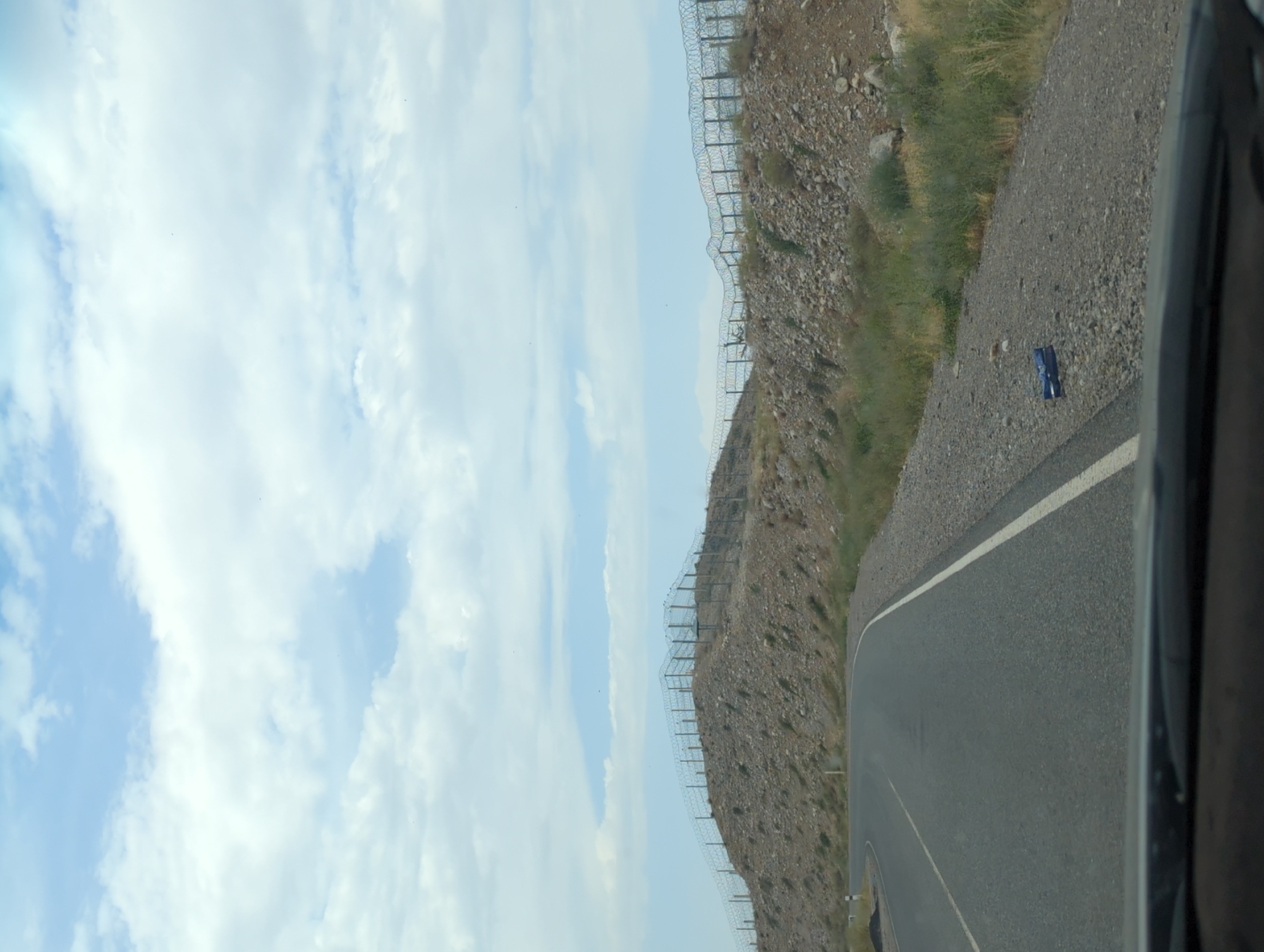 The image shows a view from a vehicle dashboard looking out at a hilly landscape. A paved road with white lane markings curves to the left. On the right side of the road, there is a rocky hillside covered with sparse vegetation. A barbed wire fence with metal posts runs along the crest of the hill, appearing to demarcate a boundary. The sky is filled with scattered white clouds against a pale blue backdrop. The scene appears to be in an outdoor, possibly rural or border area, given the presence of the fence. The location is specified as Көк-Таш, Kyrgyzstan. There are no people or animals visible in the image. A small blue object is visible on the gravel shoulder of the road.