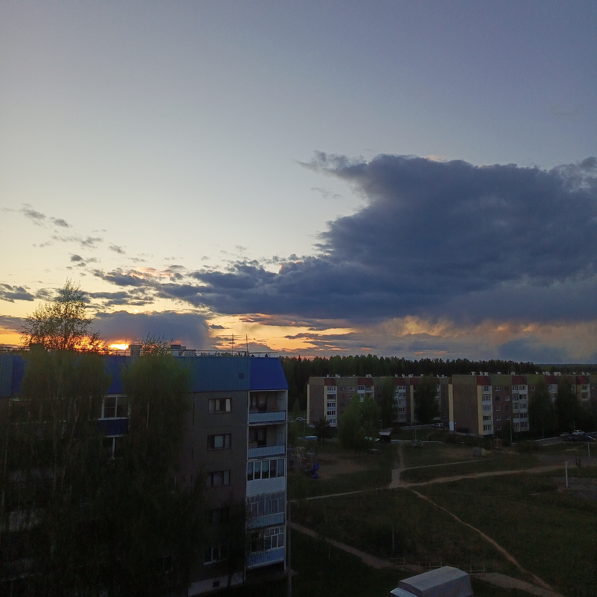 The image shows an urban landscape at sunset in Марковский, Russia. In the foreground, apartment buildings, mostly five-story, with gray facades and some blue details, are partially obscured by trees. The sky is filled with clouds ranging from light gray to dark blue, with streaks of orange light from the setting sun peeking through near the horizon. Beyond the buildings, a line of trees stretches across the middle ground, with more residential structures visible behind. In the lower right quadrant, an open area with paths suggests a park or common ground. The scene is bathed in the warm light of the setting sun, although the foreground remains somewhat shadowy.