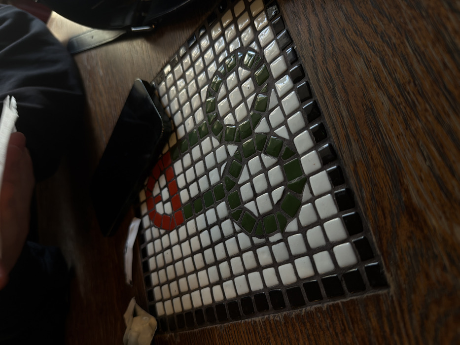The image presents an indoor scene at a wooden table in Moscow, Russia, viewed from a slightly elevated, oblique angle. A prominent square mosaic is integrated into the polished dark wood table surface. The mosaic depicts a stylized snake with a dark green body and a red head/tongue, constructed from ceramic tiles. The snake design is set against a background of white ceramic tiles, and the entire mosaic is bordered by a single row of black ceramic tiles.
On the wooden surface of the table, to the upper right of the mosaic, a dark-colored smartphone rests face down. To the upper left, a crumpled white paper napkin is visible. A person's hand, wearing dark clothing with a visible sleeve, is partially seen in the upper left portion of the frame, holding a piece of white paper. A dark strap, consistent with that of a bag or purse, is visible on the far right edge of the table.