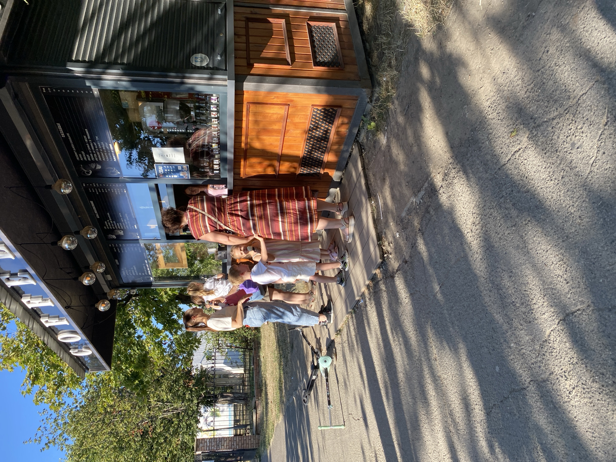 A street-level outdoor coffee kiosk, "COFFEE," is pictured in Chișinău, Moldova. The kiosk features a dark awning with hanging Edison-style light bulbs, a glass serving window displaying a menu board and bottled syrups, and a wooden base. A group of people, including two women and three children, are gathered at the serving window. One woman is ordering or receiving items, while the children appear to be waiting. One child is holding an ice cream cone. Two scooters are lying on the paved area in front of the kiosk. The scene is illuminated by bright sunlight, casting long shadows on the ground. In the background, there are trees, a fence, and a brick wall.