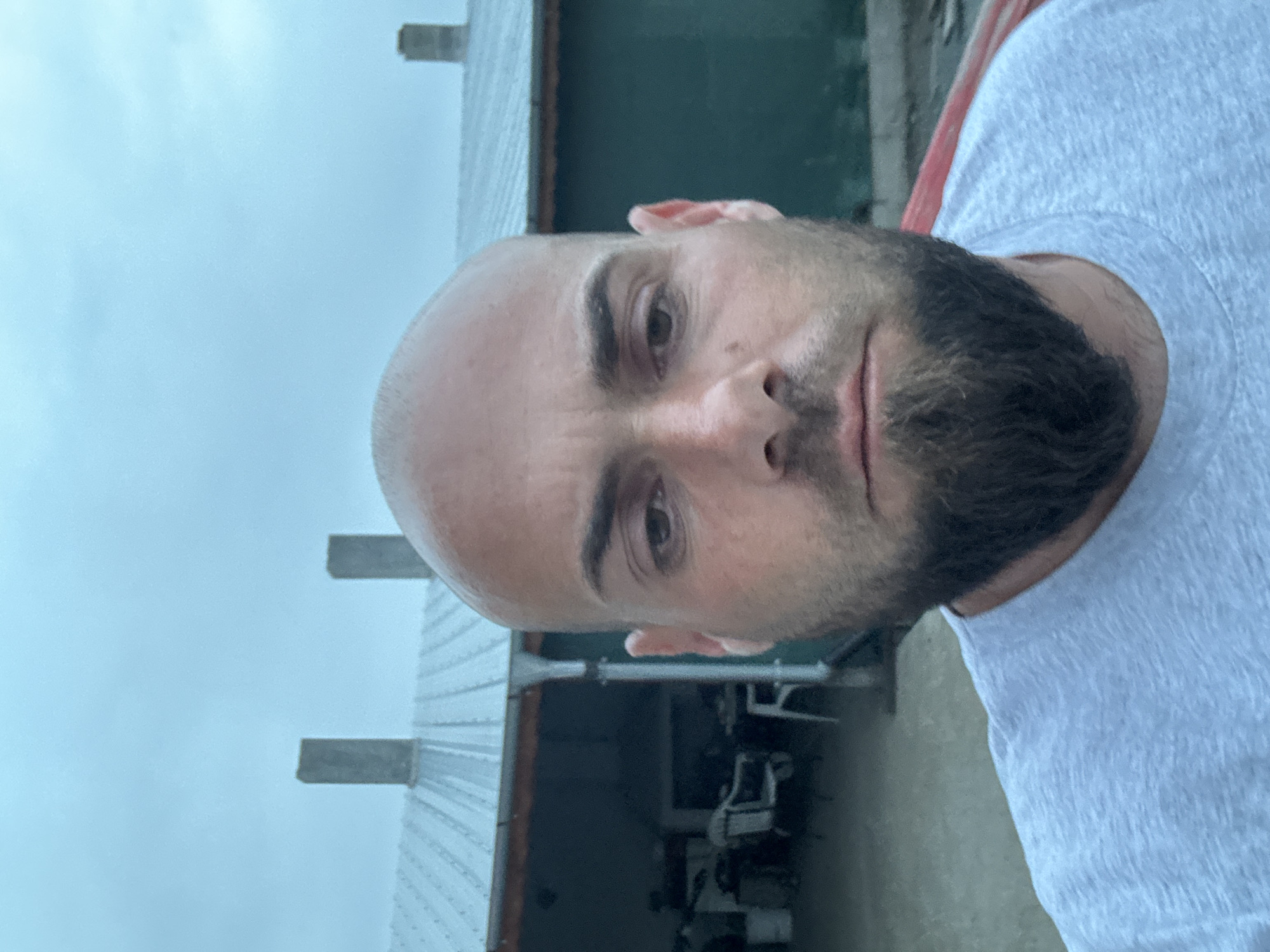 A man with a shaved head and a beard is facing the camera, outdoors in what appears to be Sarnevo, Bulgaria, based on the visual characteristics of the buildings and sky. He is wearing a light-colored t-shirt. Behind him is a building structure with multiple support beams, likely a shed or garage. The sky is overcast.