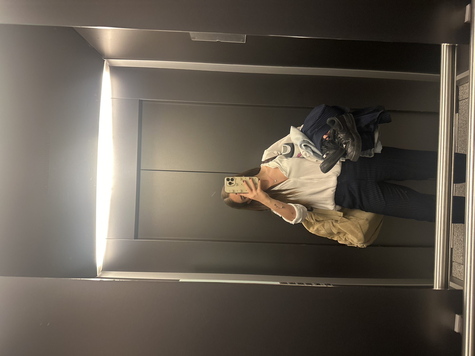 A young woman takes a mirror selfie in an elevator. She holds a phone in her right hand with her face obscured by the device. She wears a white shirt, dark striped pants, and carries a beige backpack. She holds clothes in her left arm, including what appears to be a pair of dark sneakers and a denim shirt. The interior of the elevator is gray with vertical, stainless steel handrails. The precise location in Huixquilucan de Degollado, Mexico is not discernable from the image.