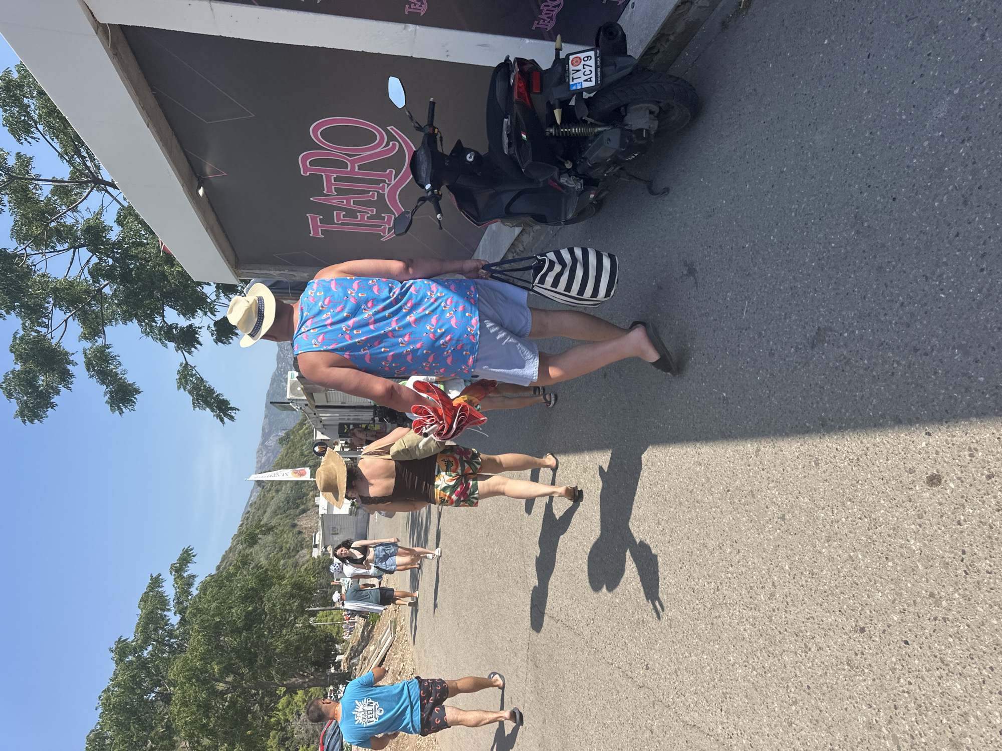 The image depicts a sunny outdoor scene, likely a pedestrian walkway in Budva, Montenegro, near a venue named "TEATRO." The foreground is dominated by a man walking away from the camera, wearing a blue tank top with a flamingo pattern and light blue shorts, and a straw hat. He is carrying a black and white striped tote bag in his left hand. To his right, a dark-colored scooter is parked next to a building with "TEATRO" written in pink lettering on its facade. The scooter has a license plate that reads "TV AC79".
Behind the man in the foreground, a woman is walking away, wearing a straw hat, a brown tank top, and patterned shorts. Further back, other individuals are seen walking, some of whom appear to be tourists. The environment suggests a warm, possibly summer day, with lush green trees lining the left side of the pathway and mountains visible in the background. The pathway itself is paved with asphalt. Shadows are cast on the ground, indicating the direction of the sun. The overall impression is that of a lively tourist area.