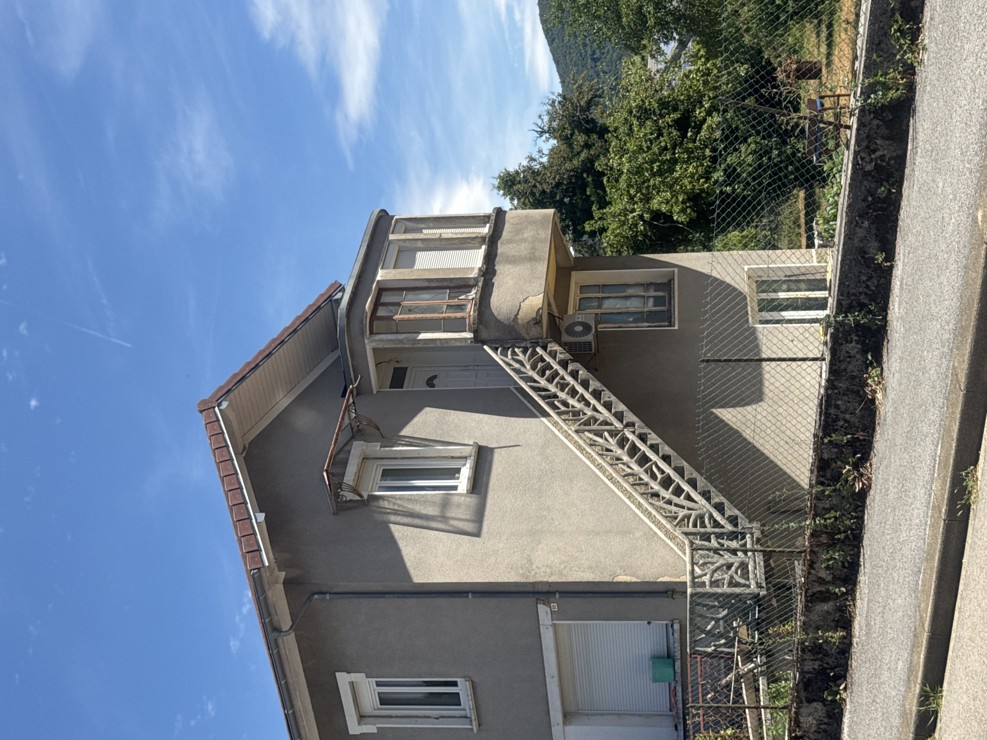 The image shows the exterior of a grey, two-story house with a brown tiled roof, located in Coteaux du Lizon, France. The house has several windows, some with white frames and blinds. An external staircase with a decorative metal railing leads to the upper floor. A small balcony or bay window protrudes from the upper front of the house. An air conditioning unit is mounted on the exterior wall below the balcony. The house is surrounded by a chain-link fence, with greenery and trees visible behind it, and a hilly, forested landscape in the background. A paved road and curb are in the foreground. The sky is blue with some scattered white clouds.