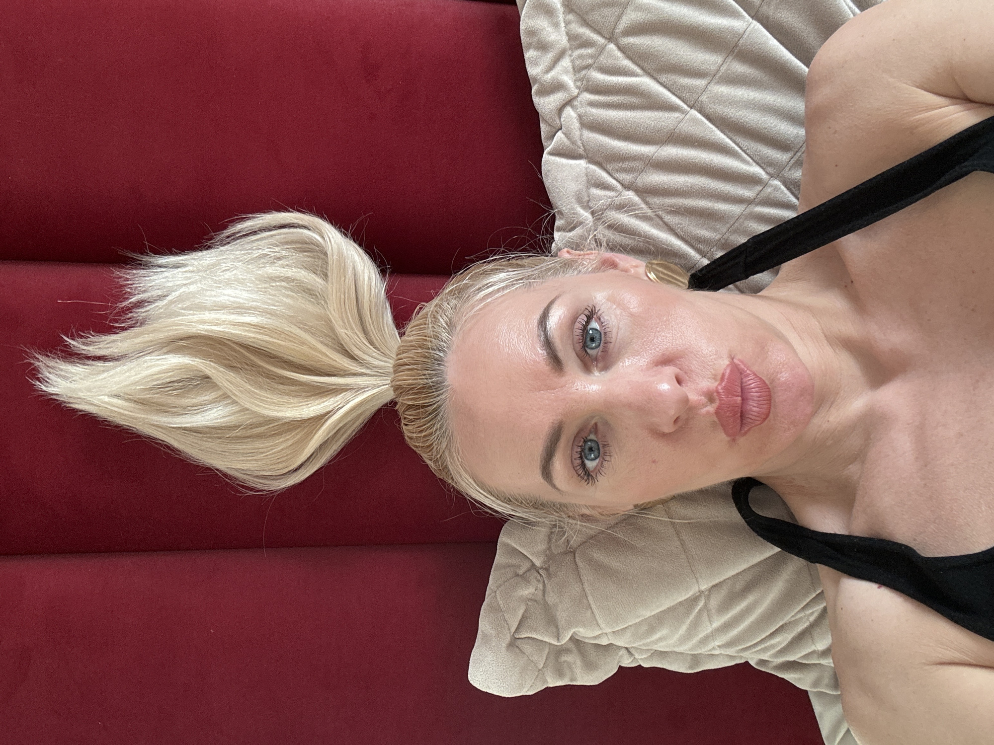 A woman lies on a red surface and a beige pillow. She has blonde hair tied in a high ponytail and blue eyes. She wears a black top. The location is Unknown, Unknown.
